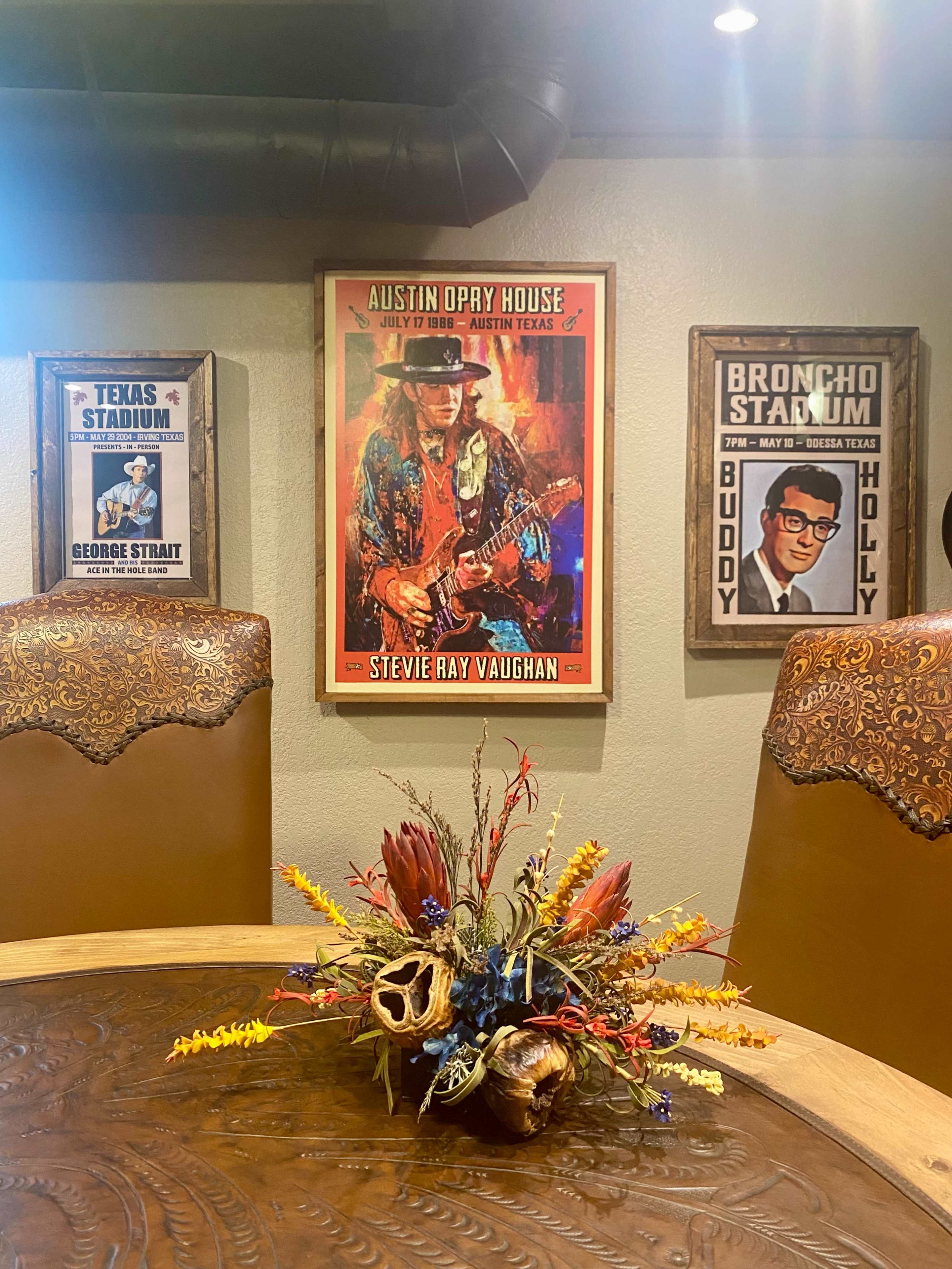A dining area with a circular wooden table, decorated with a floral arrangement, and walls featuring framed posters of music venues and artists, including an Austin Opry House poster of Stevie Ray Vaughan.