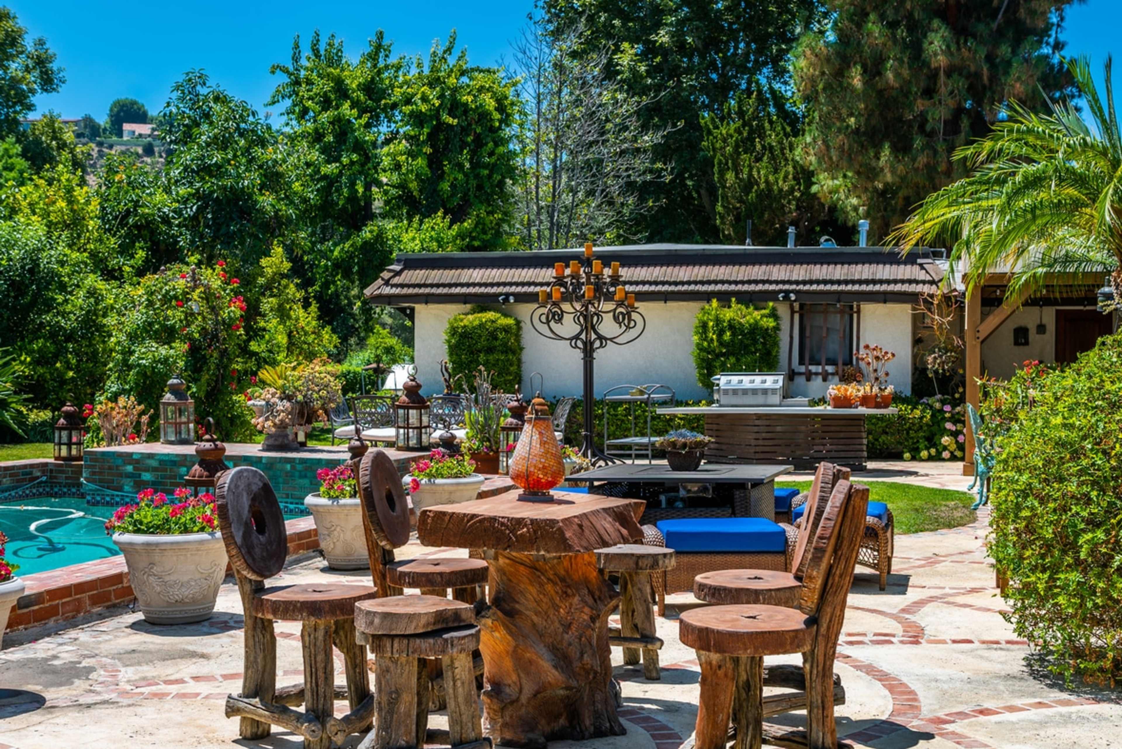 The image depicts a secluded outdoor patio area featuring a wooden table surrounded by carved stools, with a swimming pool in the background and lush greenery throughout the scene.