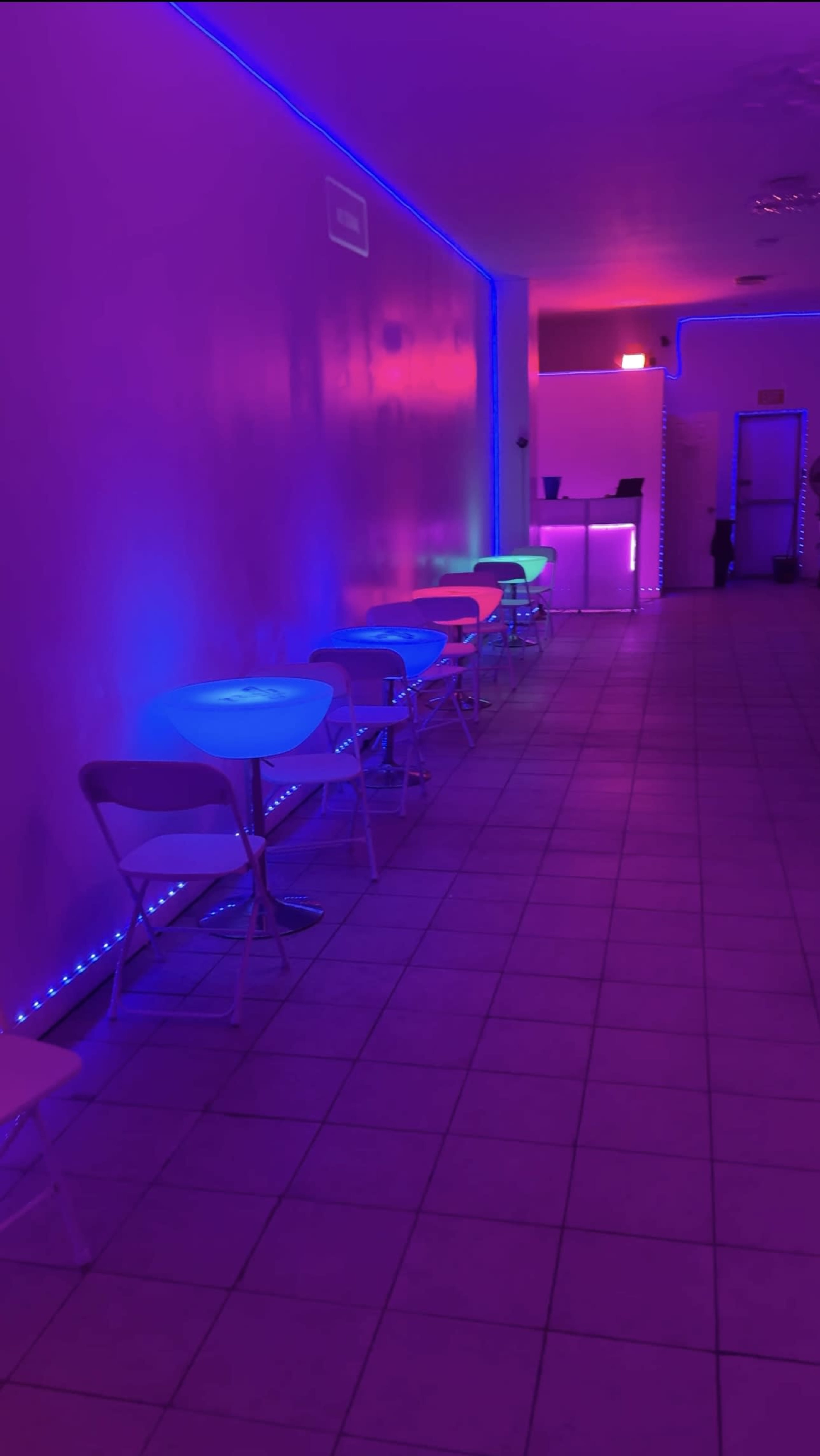 The image shows a narrow room lined with brightly colored illuminated tables and chairs against a purple and pink lit wall.