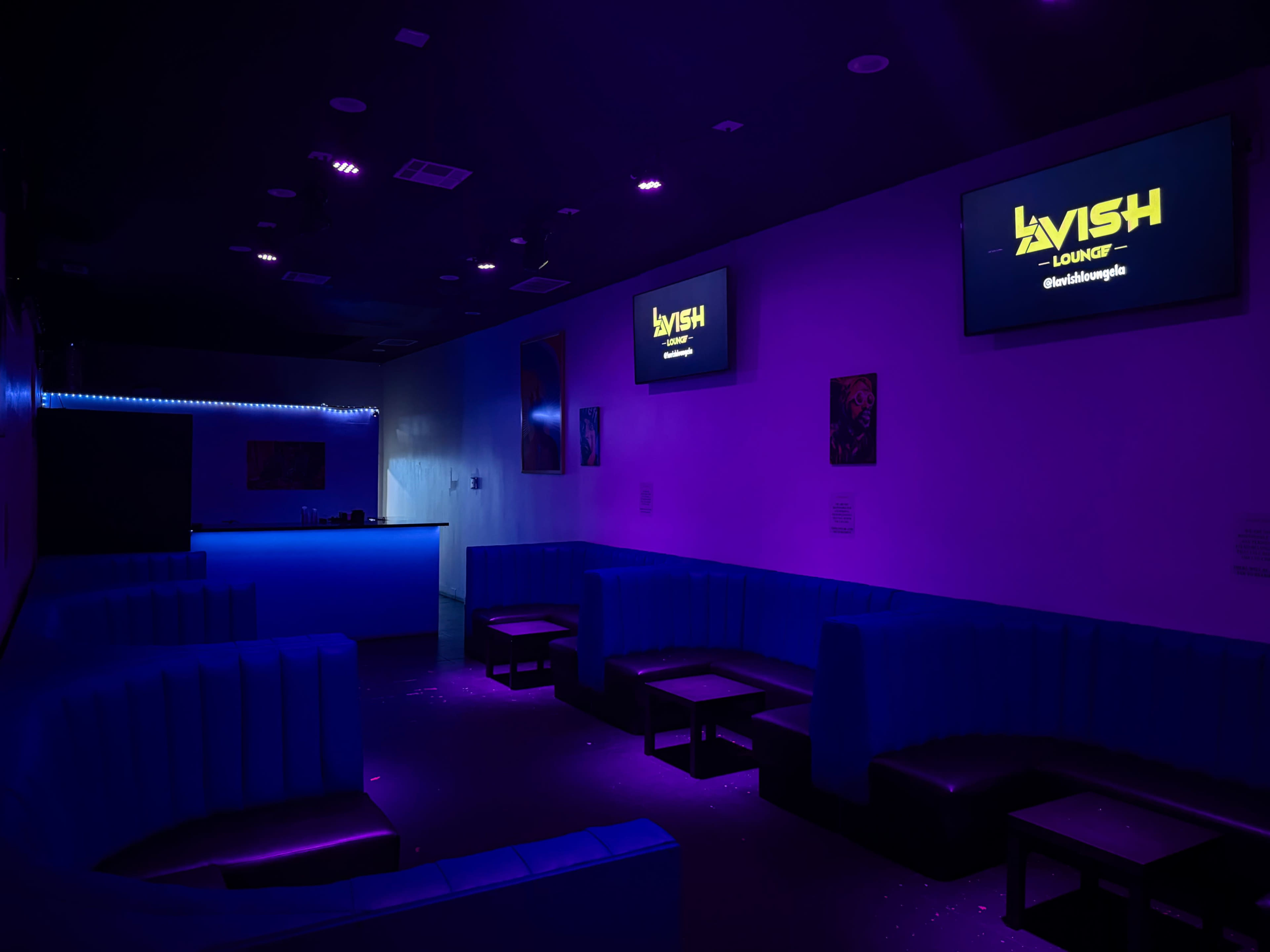 The image shows a dimly lit lounge with blue seating, illuminated by purple and blue lights, featuring two screens displaying the name "Lavish Lounge."