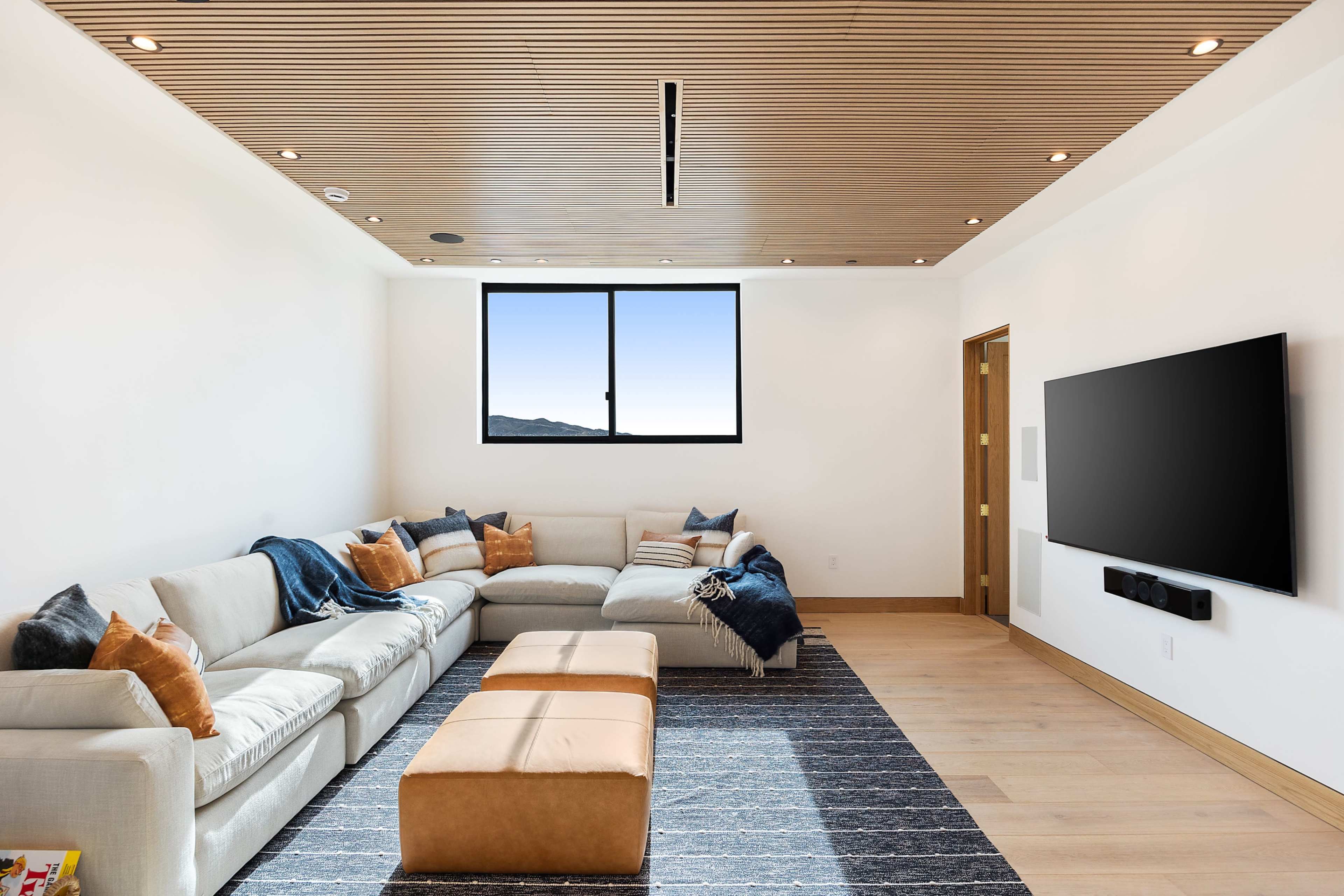 A spacious living room features a large sectional sofa, a coffee table, and a wall-mounted television, with a window showcasing a view of the outdoors.
