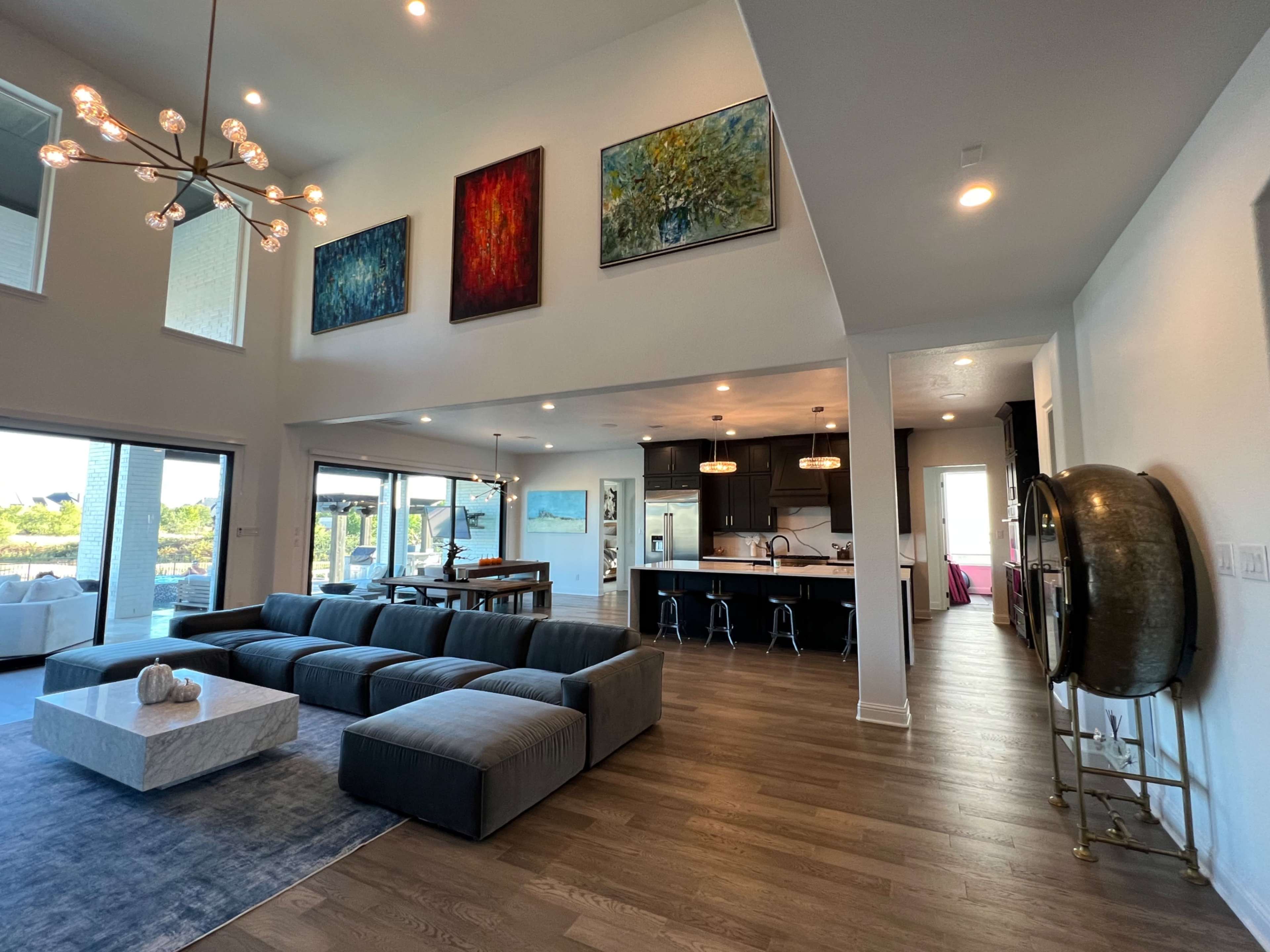 The image shows a spacious, modern living area featuring large windows, a sectional sofa, and colorful artwork hanging on the walls, with a kitchen visible in the background.