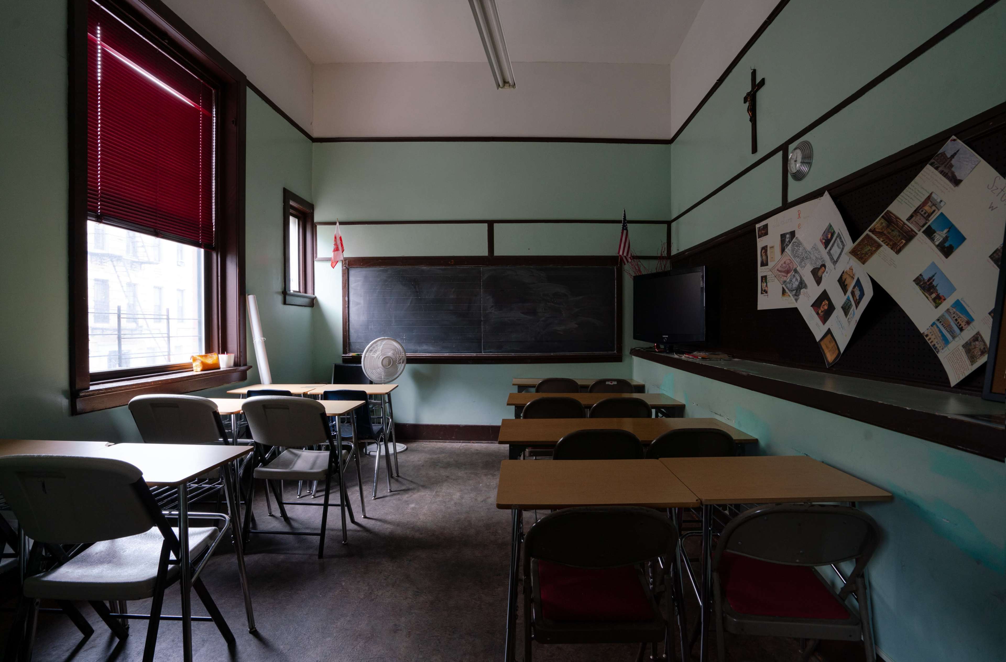 A classroom features rows of desks, a chalkboard, and a fan in a space with green walls and red blinds.
