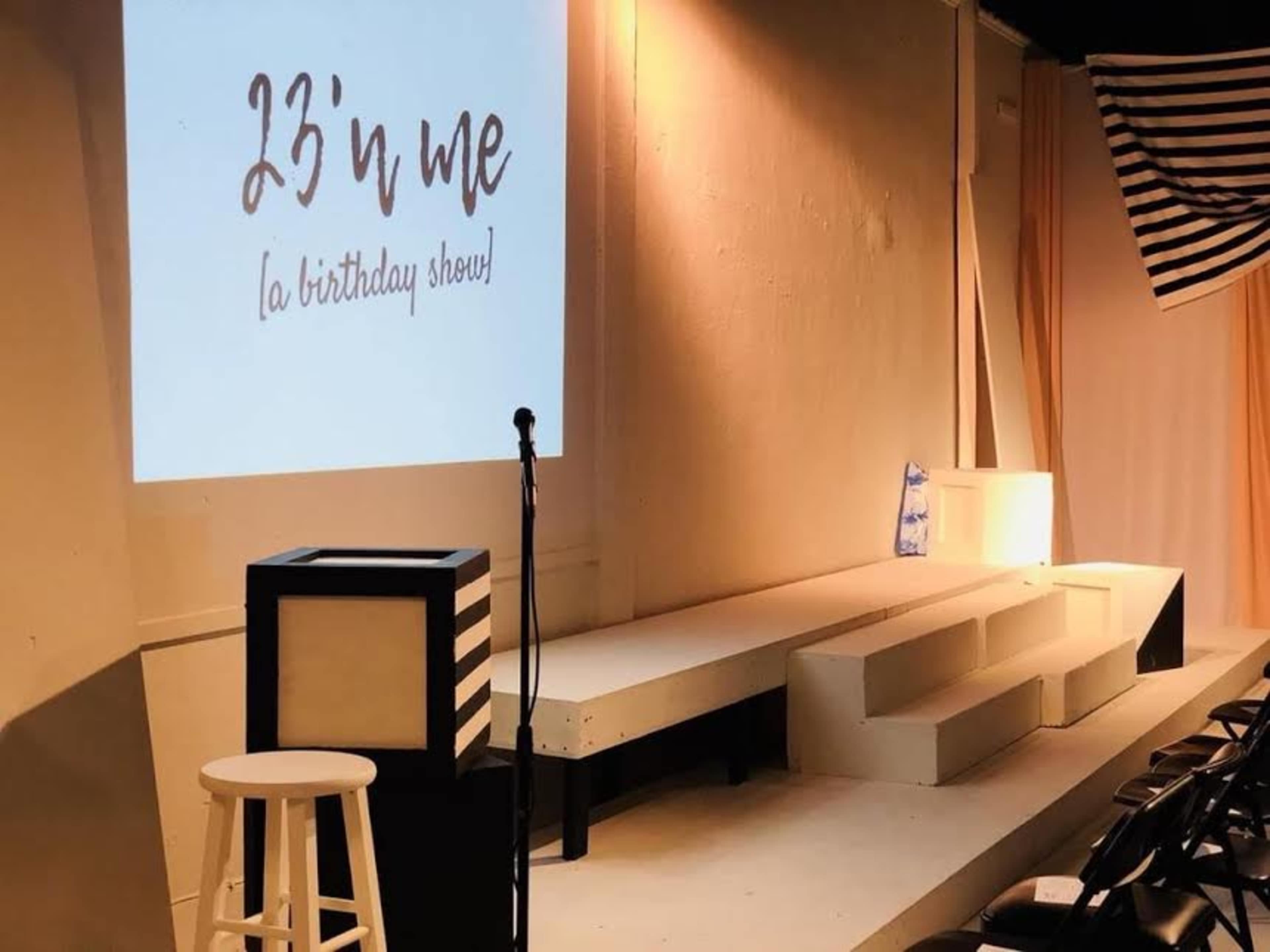 The image shows a stage setup for a birthday show titled "23 'n Me," featuring a microphone on a stand, a stool, and tiered seating.