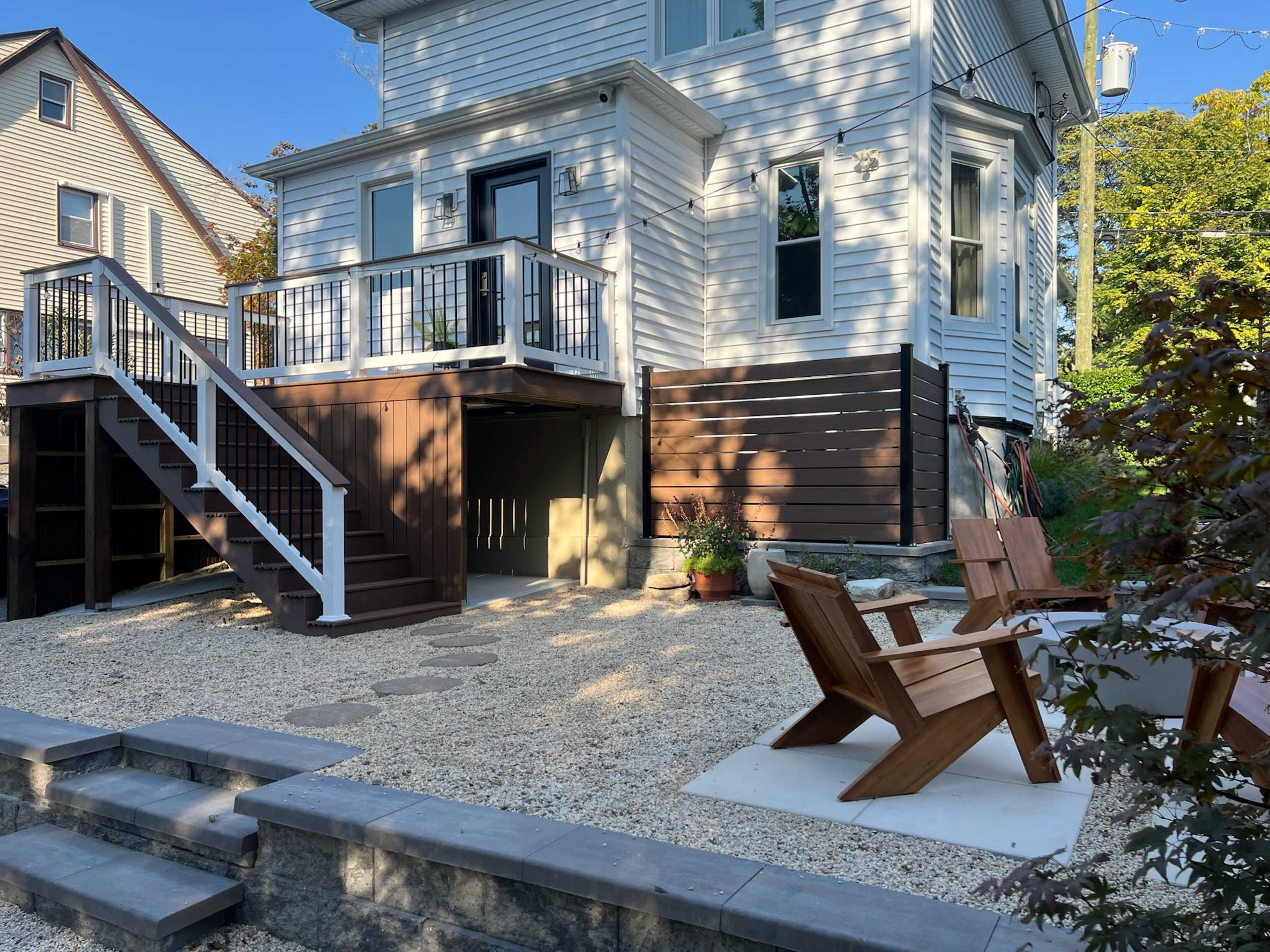 The image shows a modern two-story house with a deck and stairs leading to a gravel patio area featuring wooden chairs and stone steps.