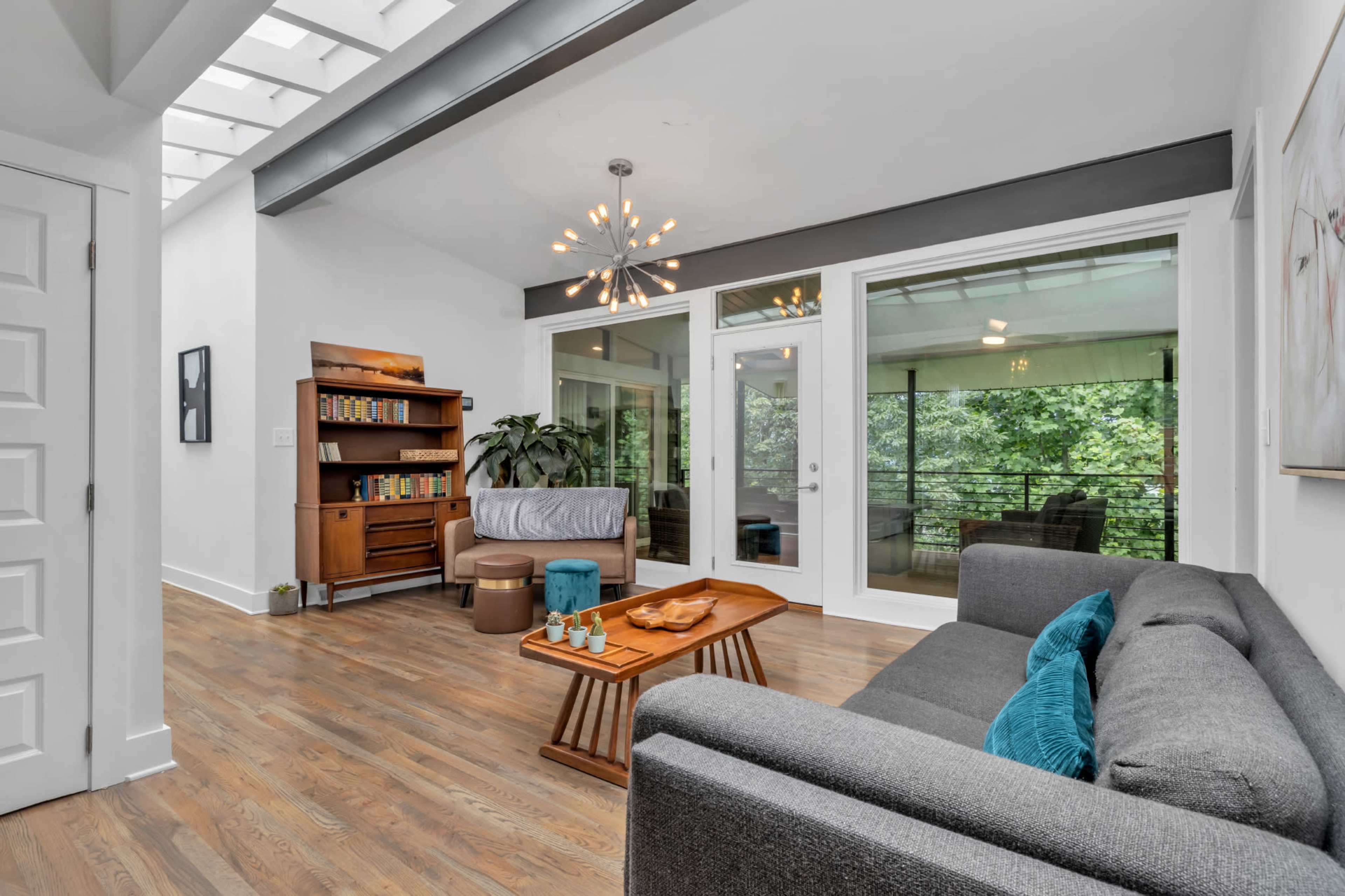 The room features a modern design with a gray sofa, a wooden coffee table, and large glass doors that lead to a balcony surrounded by greenery.