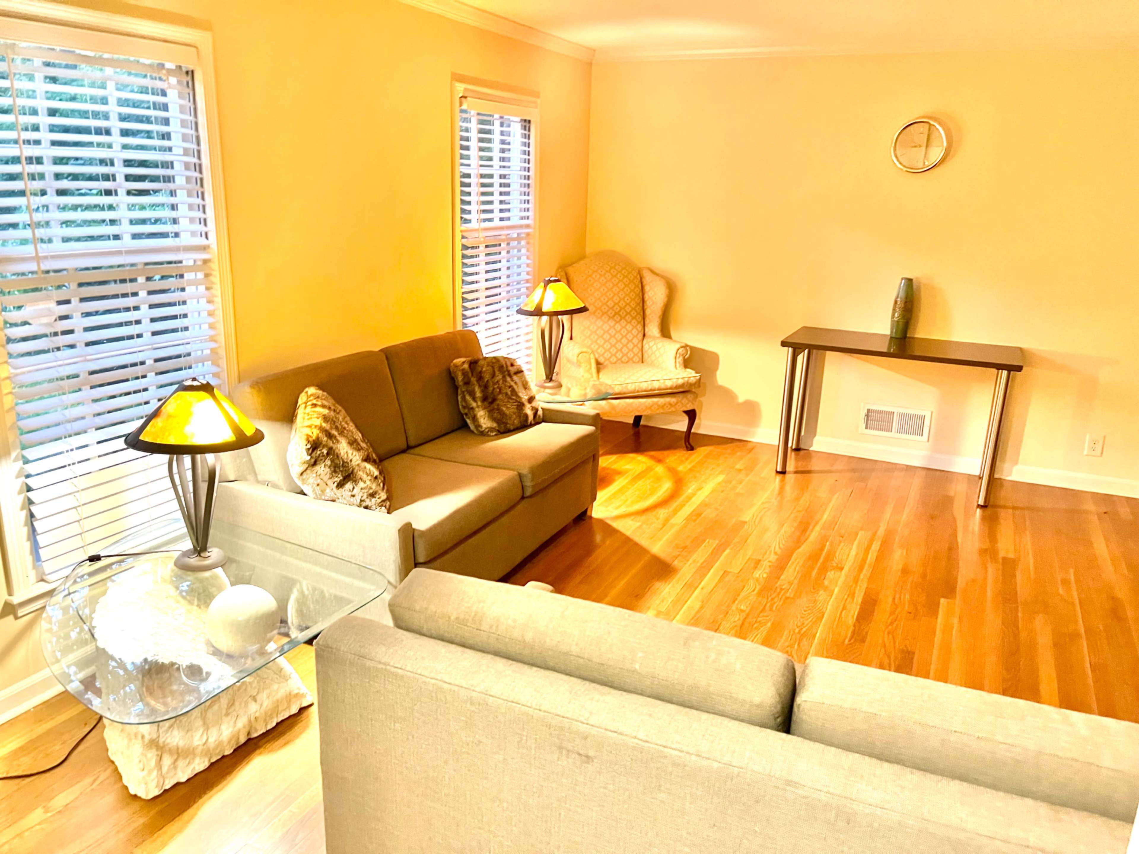 The image shows a cozy living room featuring two sofas, a chair, a small table, and a lamp, illuminated by natural light from the windows.