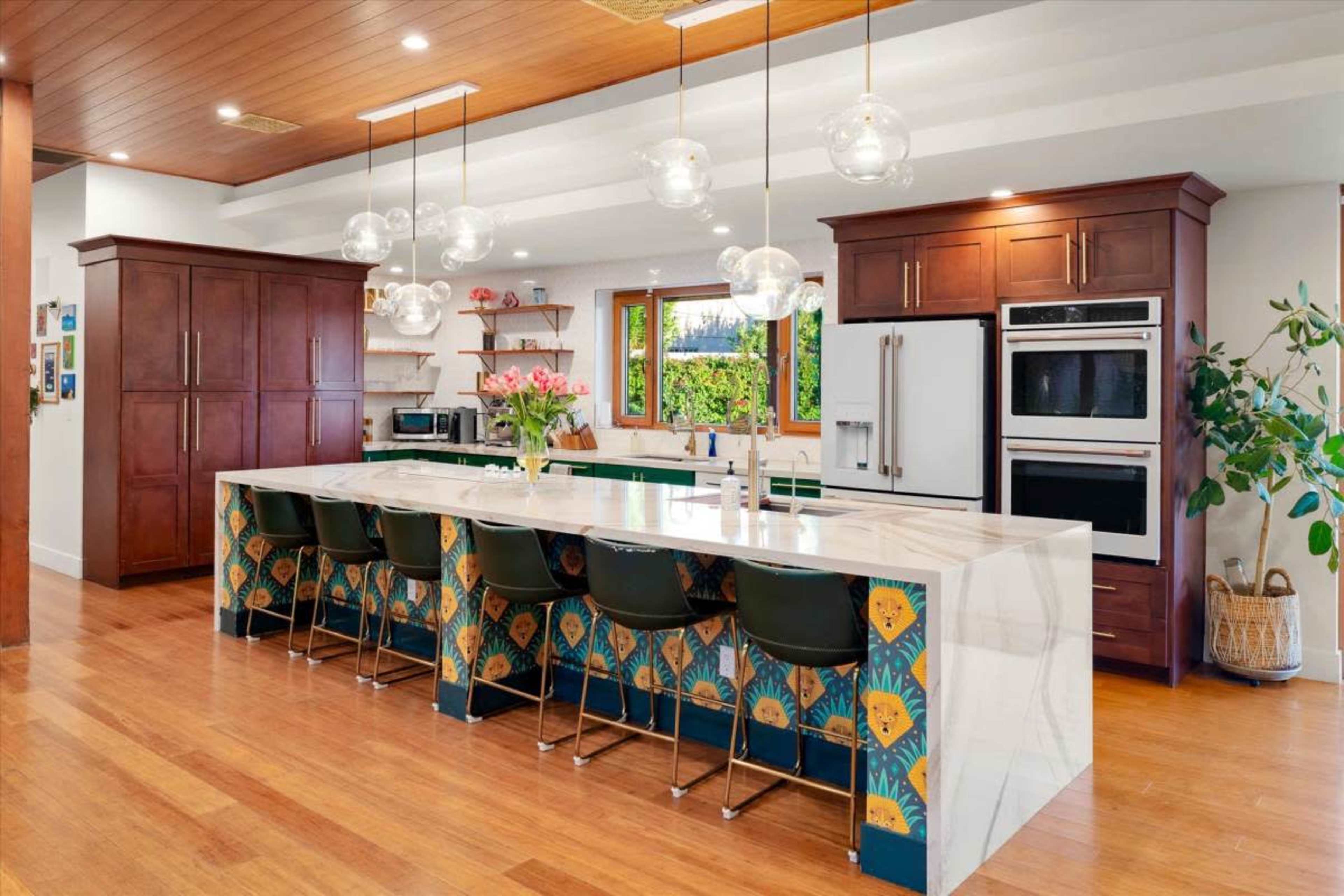 The image shows a modern kitchen with a large island featuring seating, dark wood cabinetry, and stylish light fixtures.