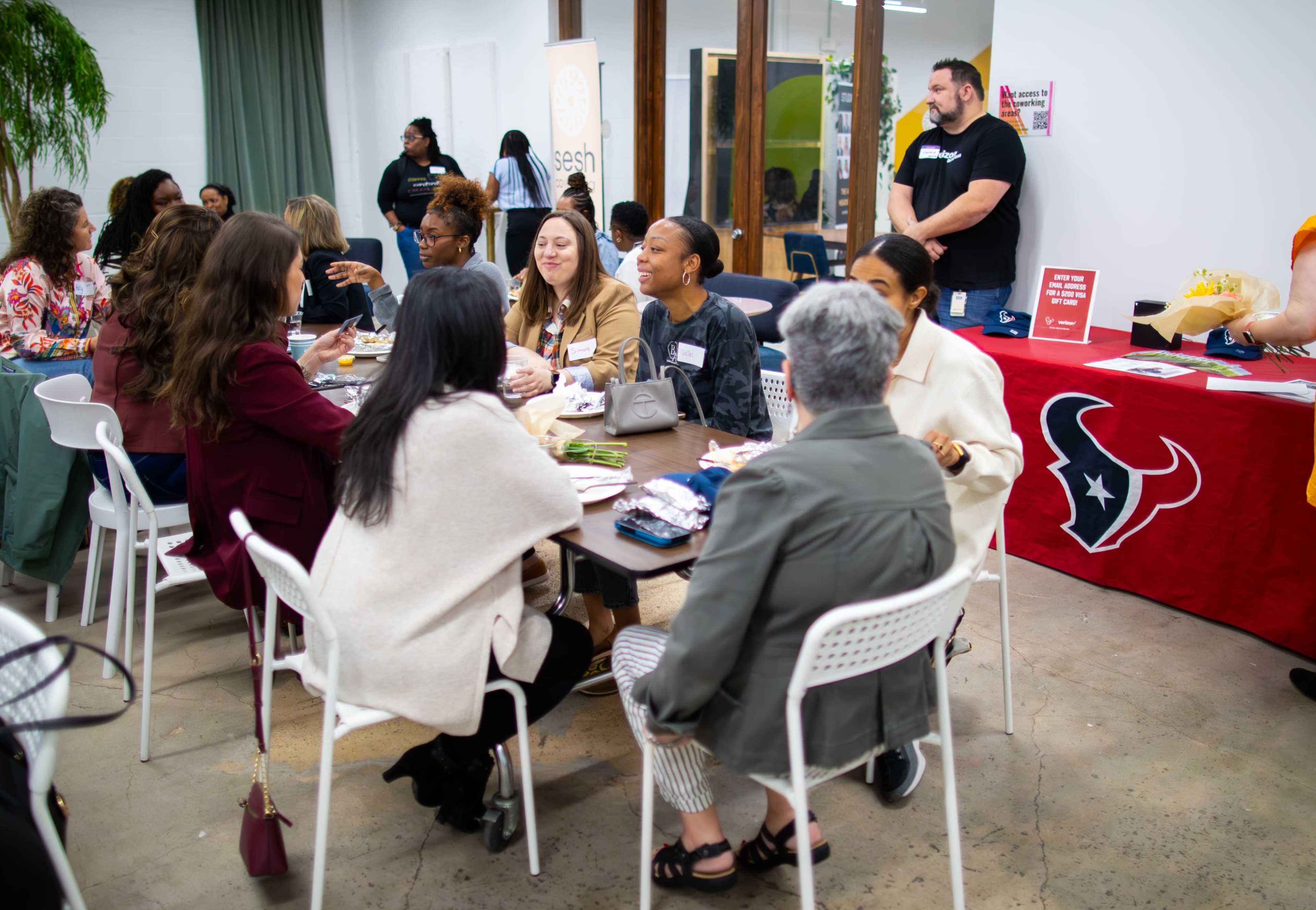 A group of women sits around a table in a casual meeting space, engaging in conversation and dining together.