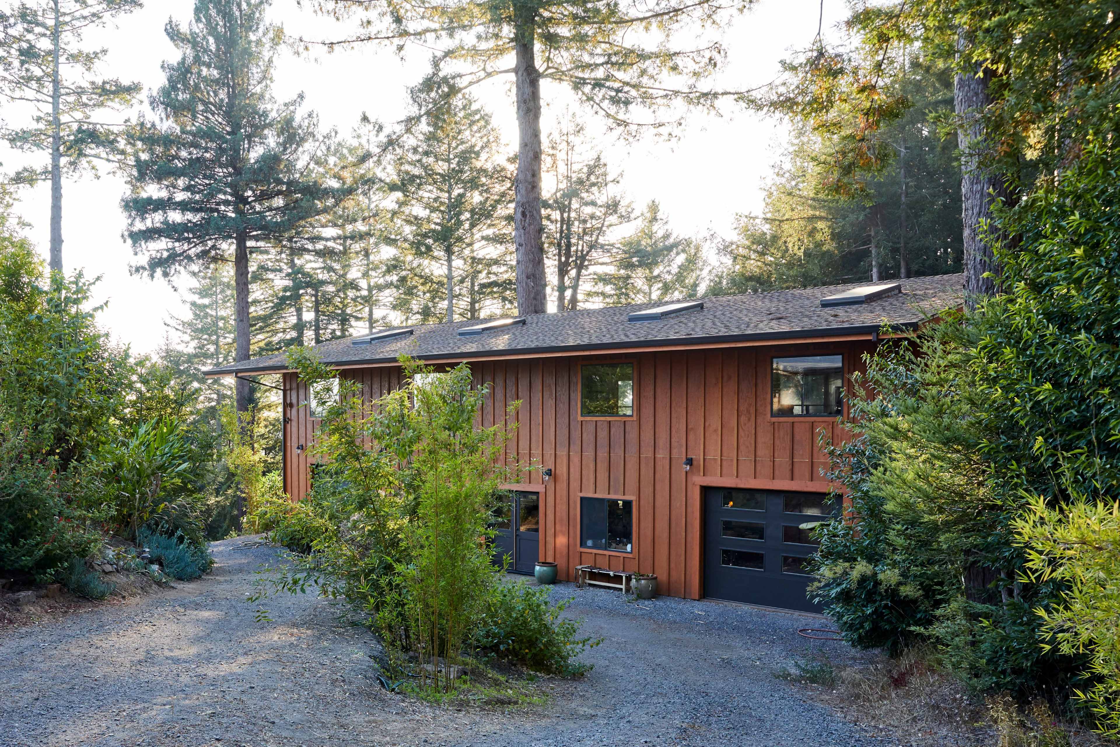 A modern wooden house is situated among tall trees with a gravel driveway leading up to it.
