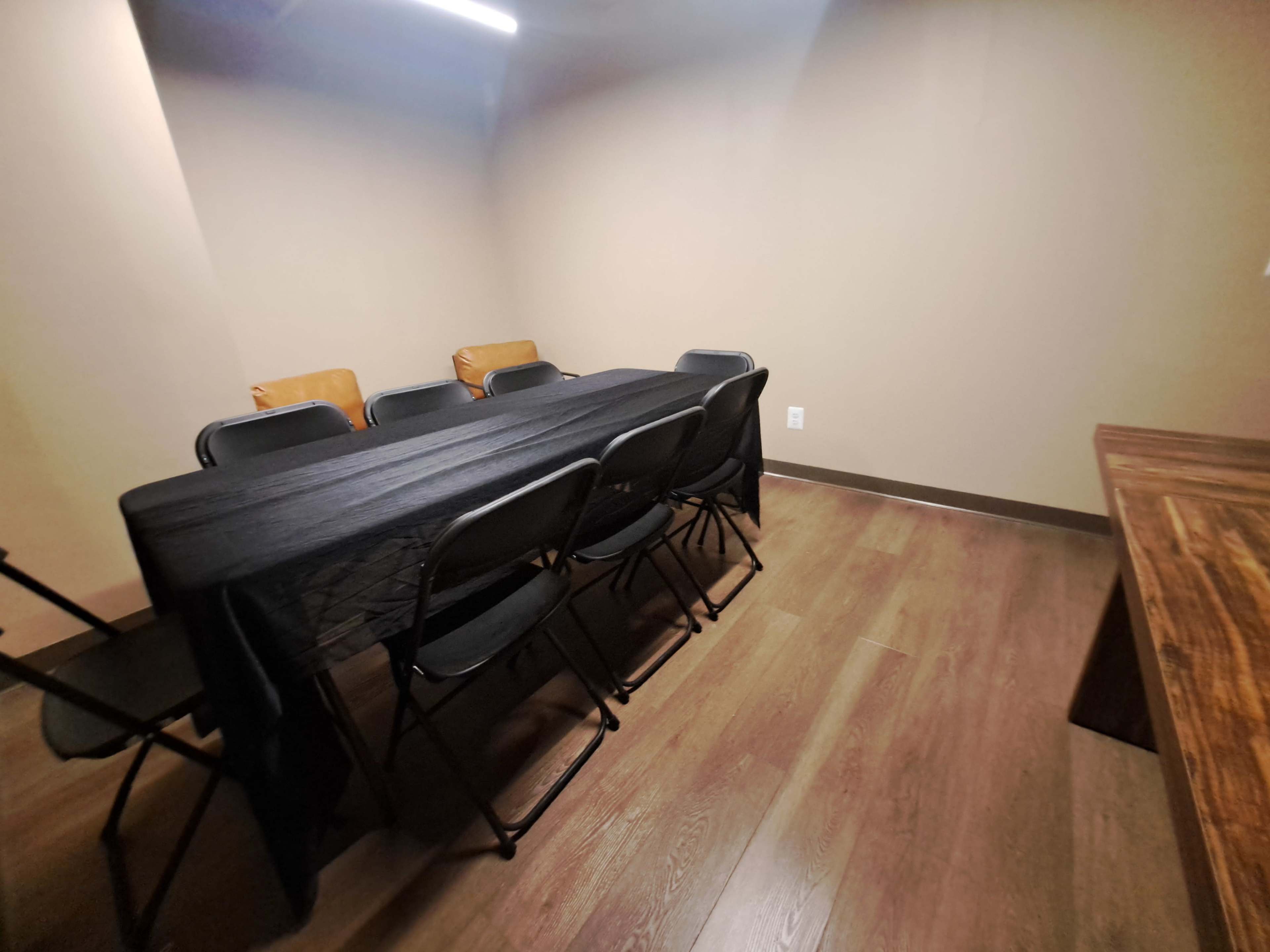 The image shows a sparsely furnished room with a long black table surrounded by black folding chairs.