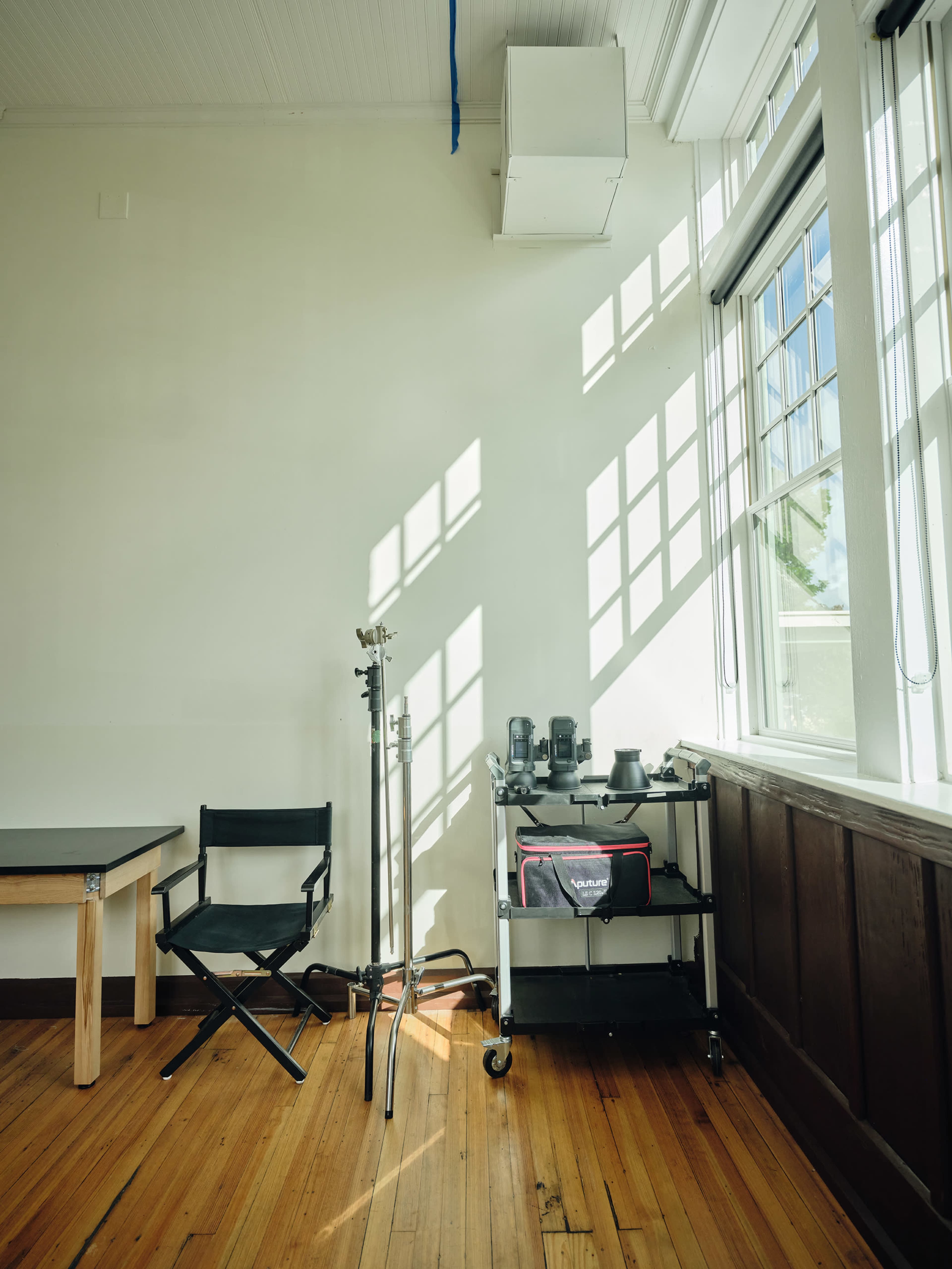 The image shows a bright, empty room with large windows casting shadows on the wooden floor, featuring a black folding chair, a stand, and a cart with camera equipment.