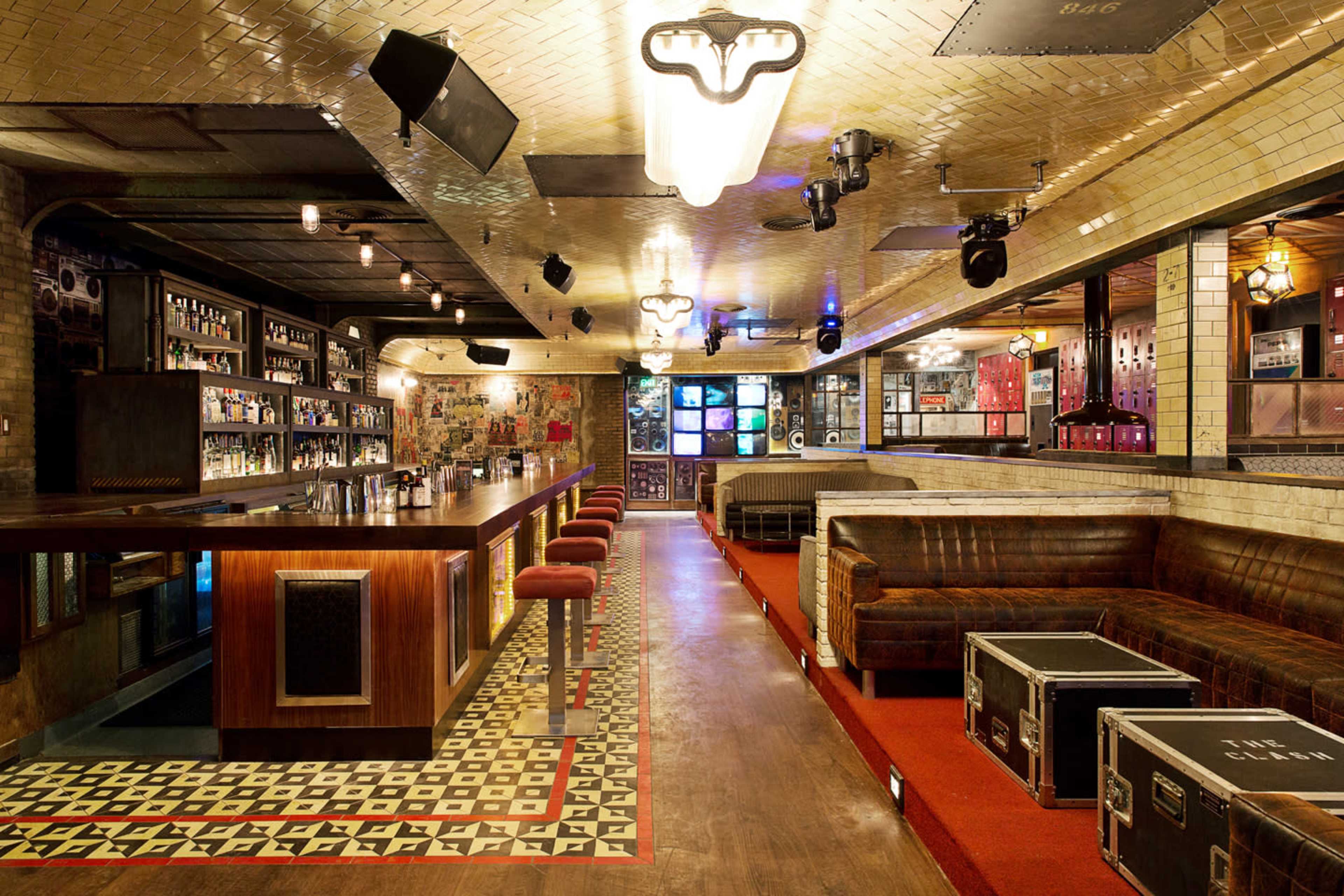 The image depicts a stylish bar interior with a long wooden bar, patterned tile flooring, and cozy seating areas along the walls.