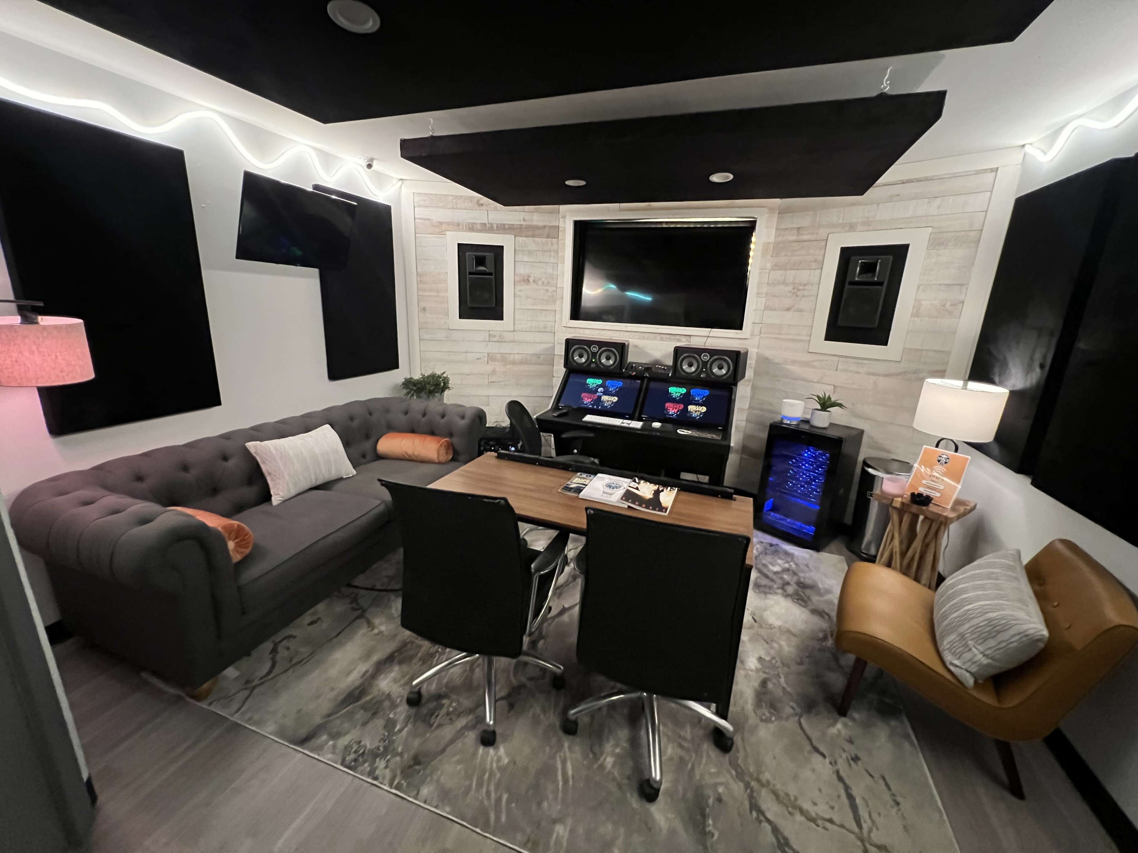 A modern recording studio with a gray sofa, a wooden table with two black chairs, and sound equipment mounted on the walls, all set against a light-colored wood panel background.