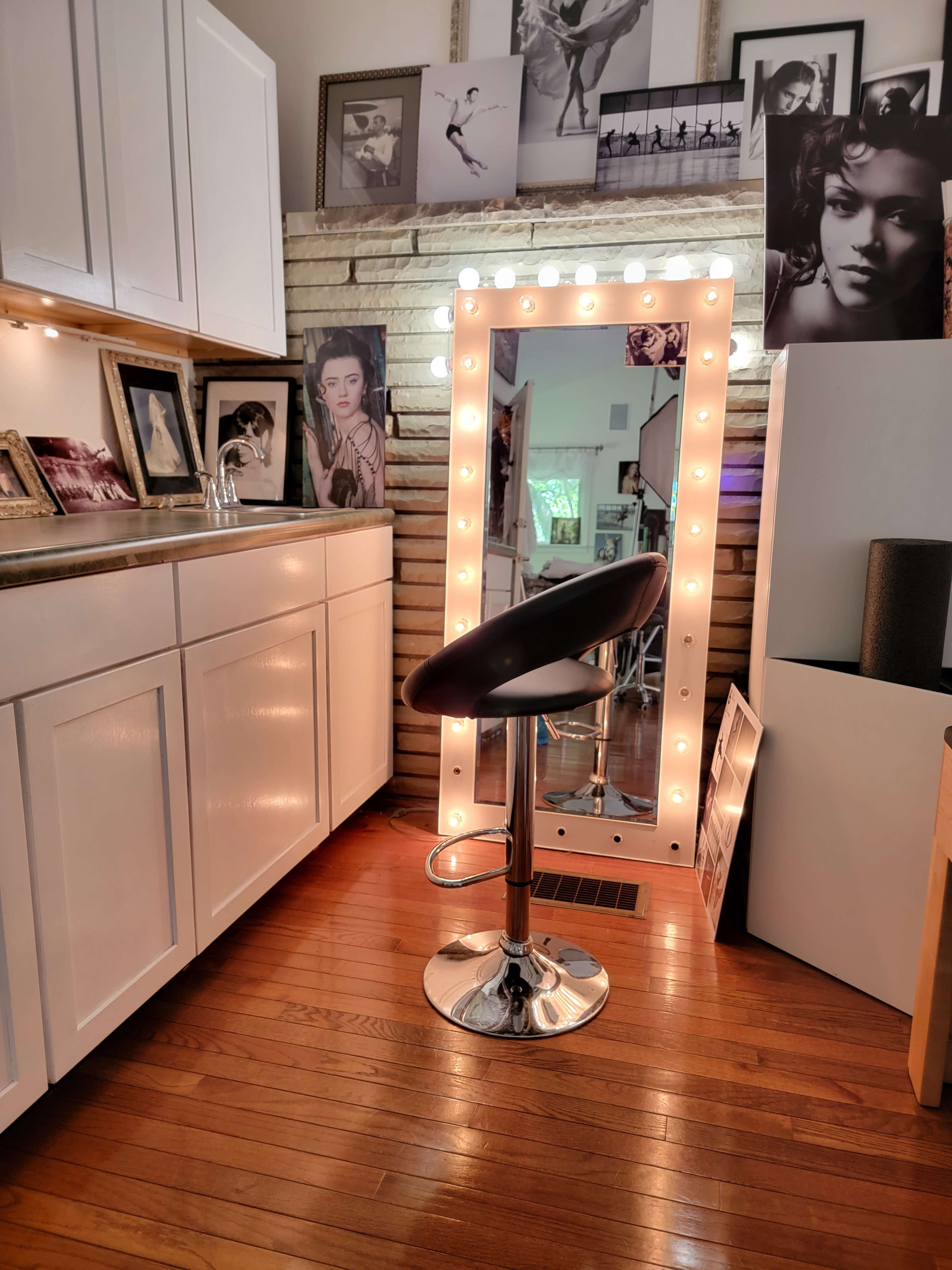 The image shows a well-lit vanity area with a large mirror framed by light bulbs, a black swivel stool, and several framed photos on the walls.