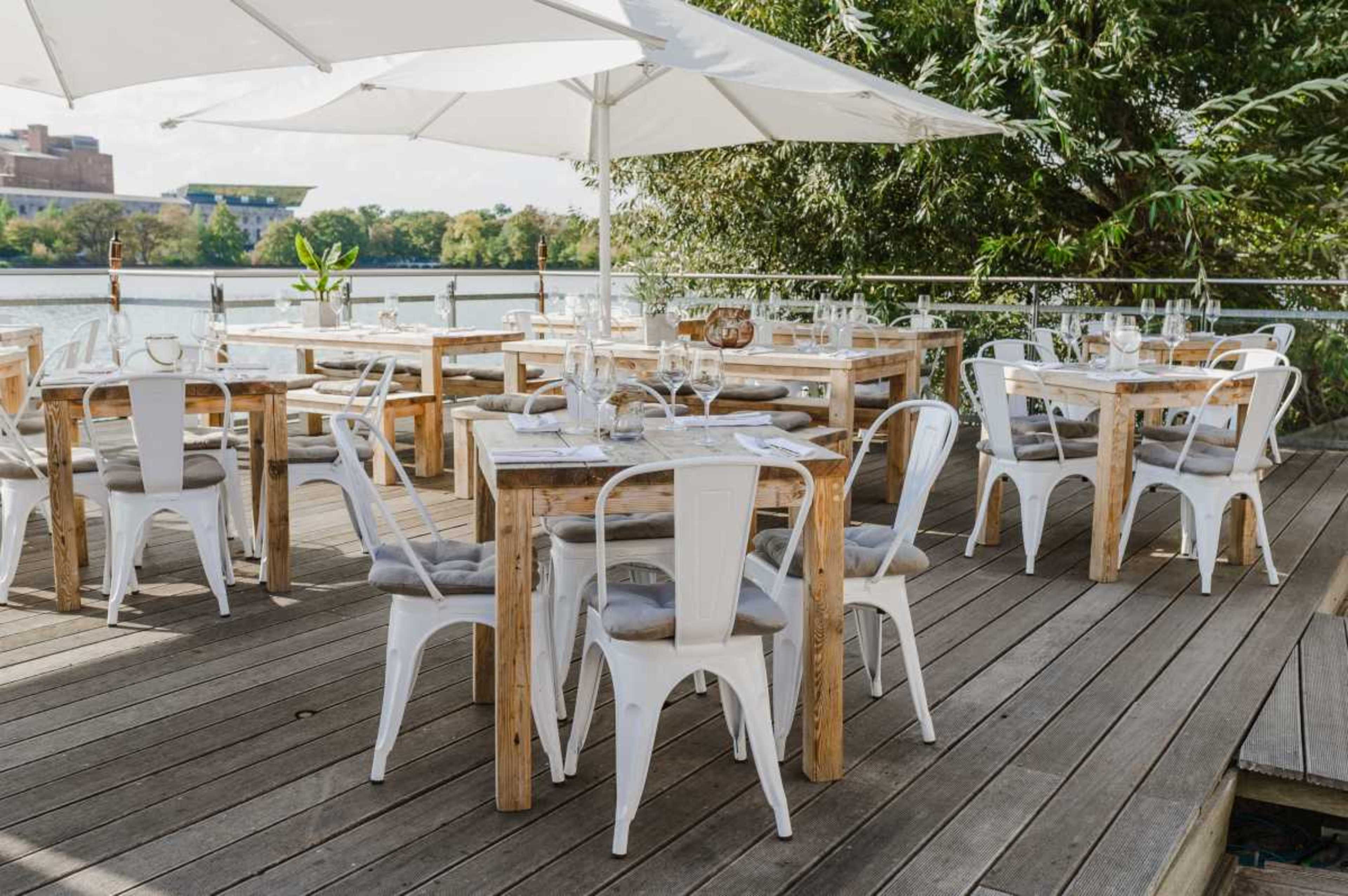 The image shows an outdoor dining area with wooden tables and metal chairs under a large white umbrella, overlooking a body of water.