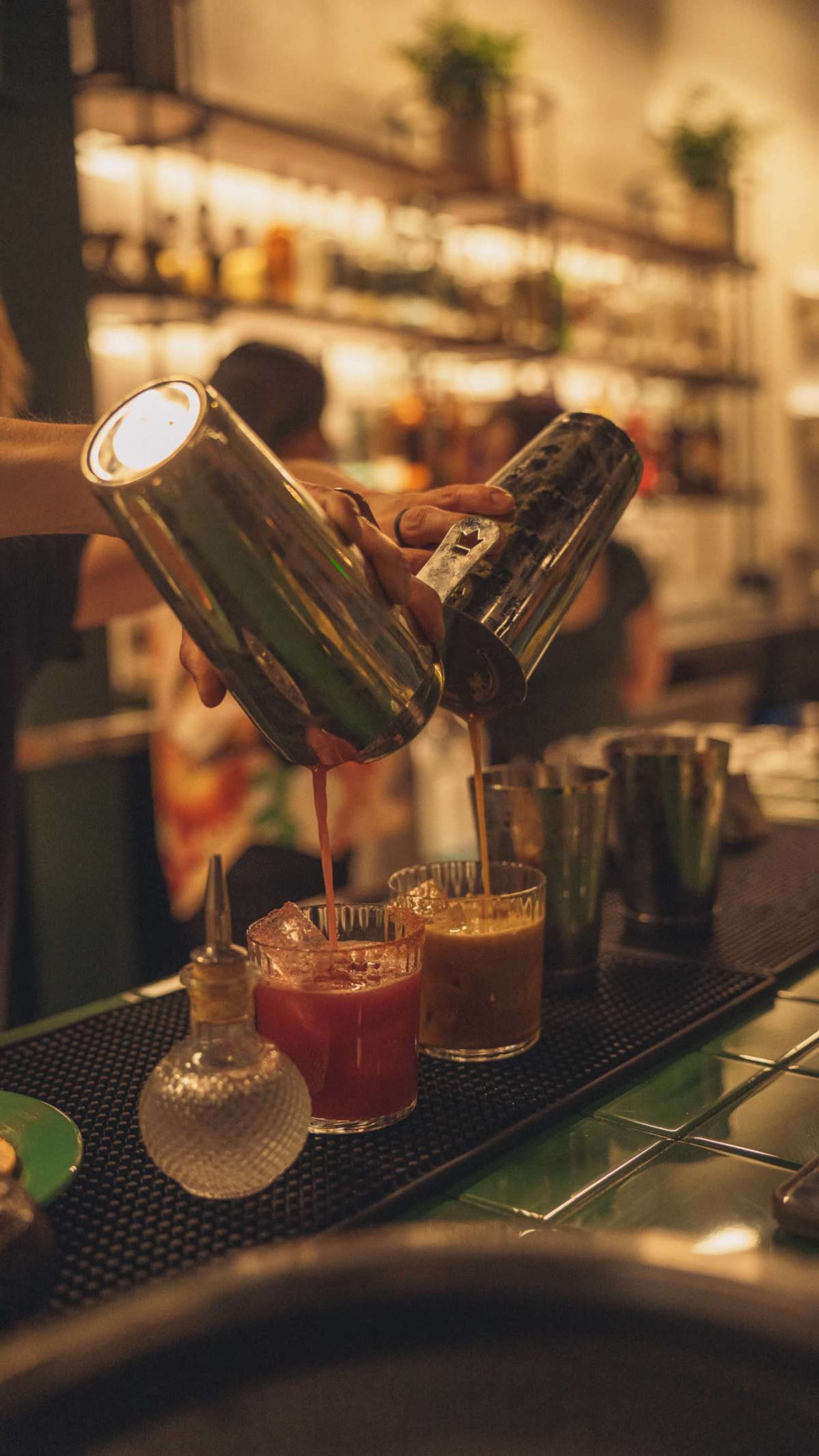 A bartender is pouring cocktails from two shakers into glasses on the bar.