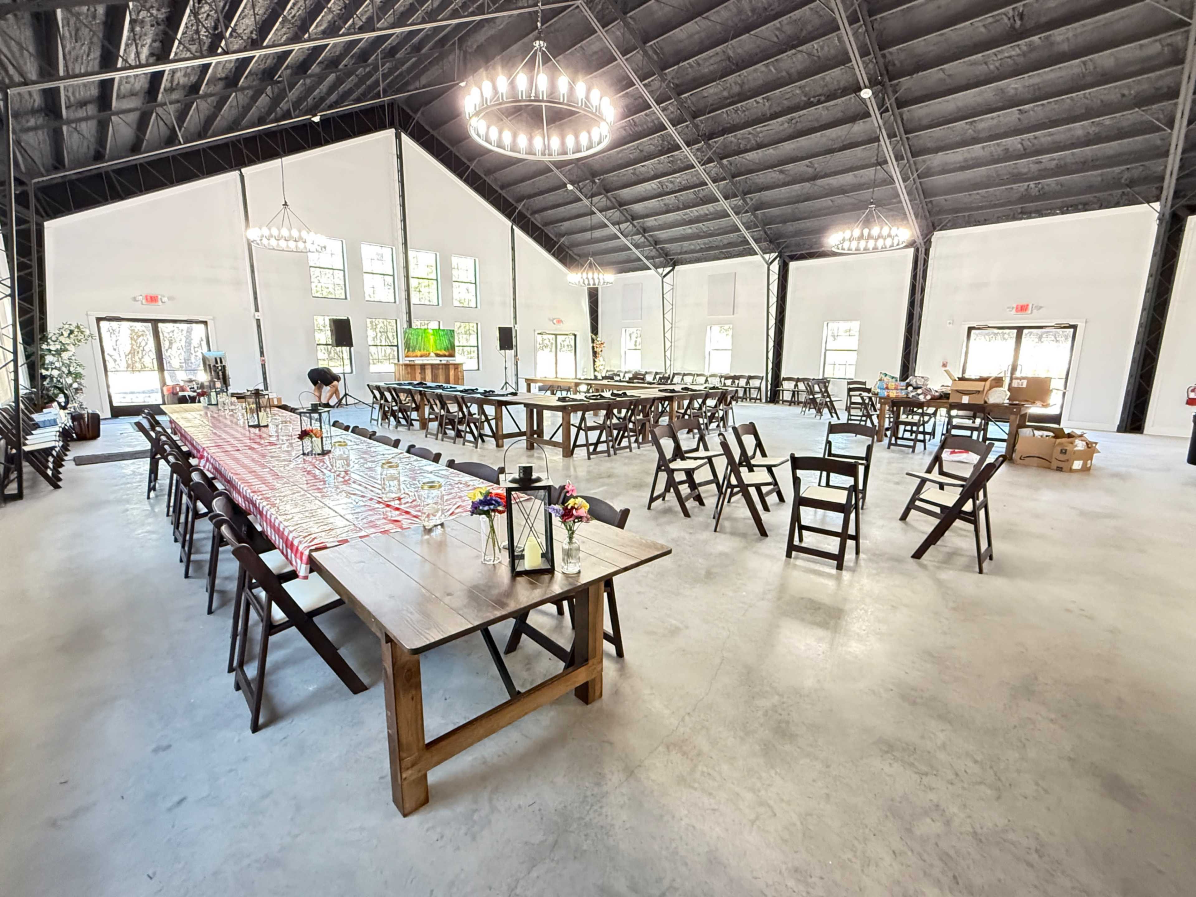 The image shows an indoor event space with wooden tables and chairs arranged for a gathering, featuring high ceilings and large windows providing natural light.
