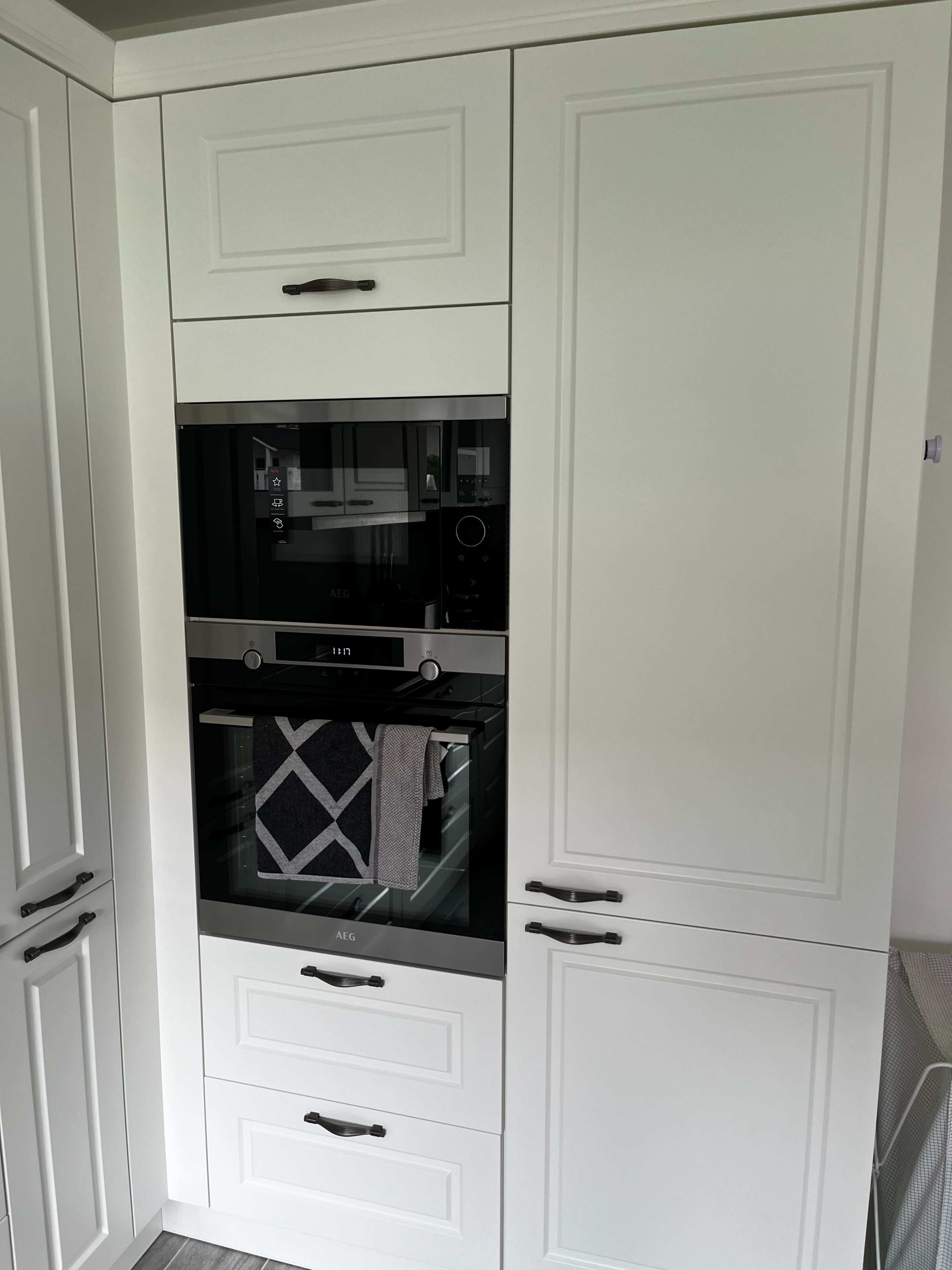 The image shows a modern kitchen cabinet with a built-in AEG oven and microwave, featuring white cabinetry and a grey towel hanging on the oven door.