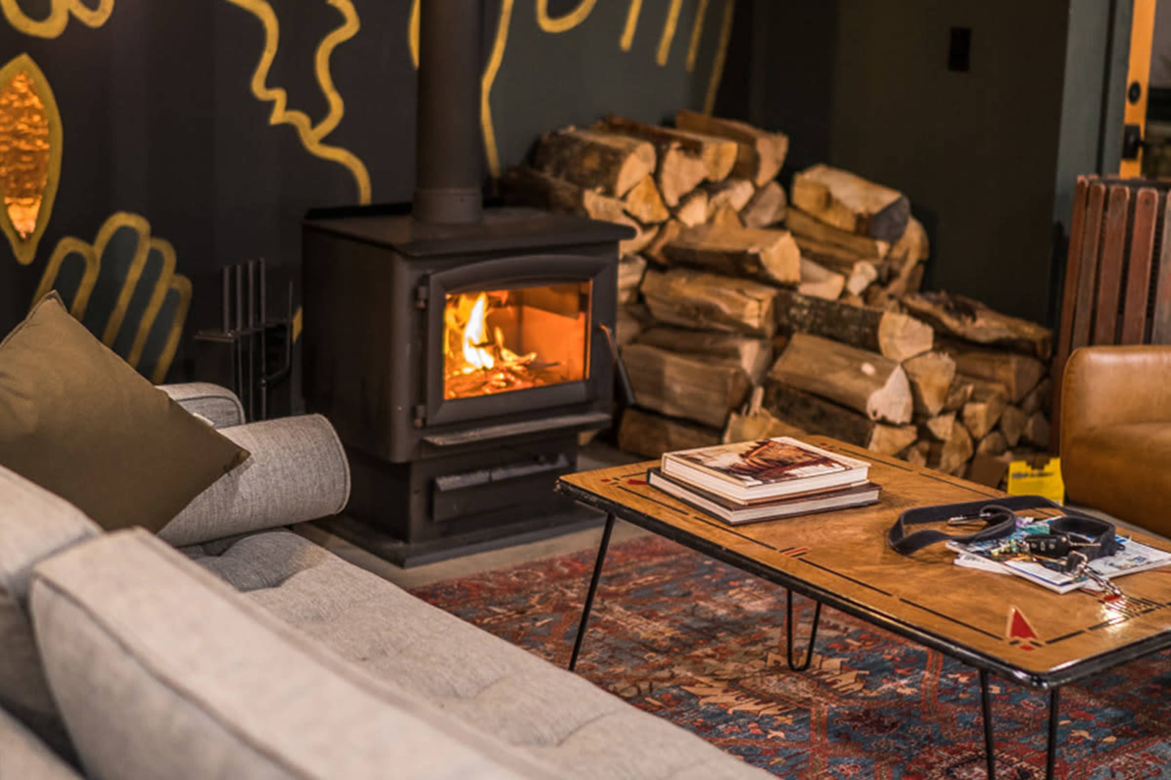 A cozy living room with a wood stove, a stack of firewood, a coffee table with magazines, and a gray sofa.