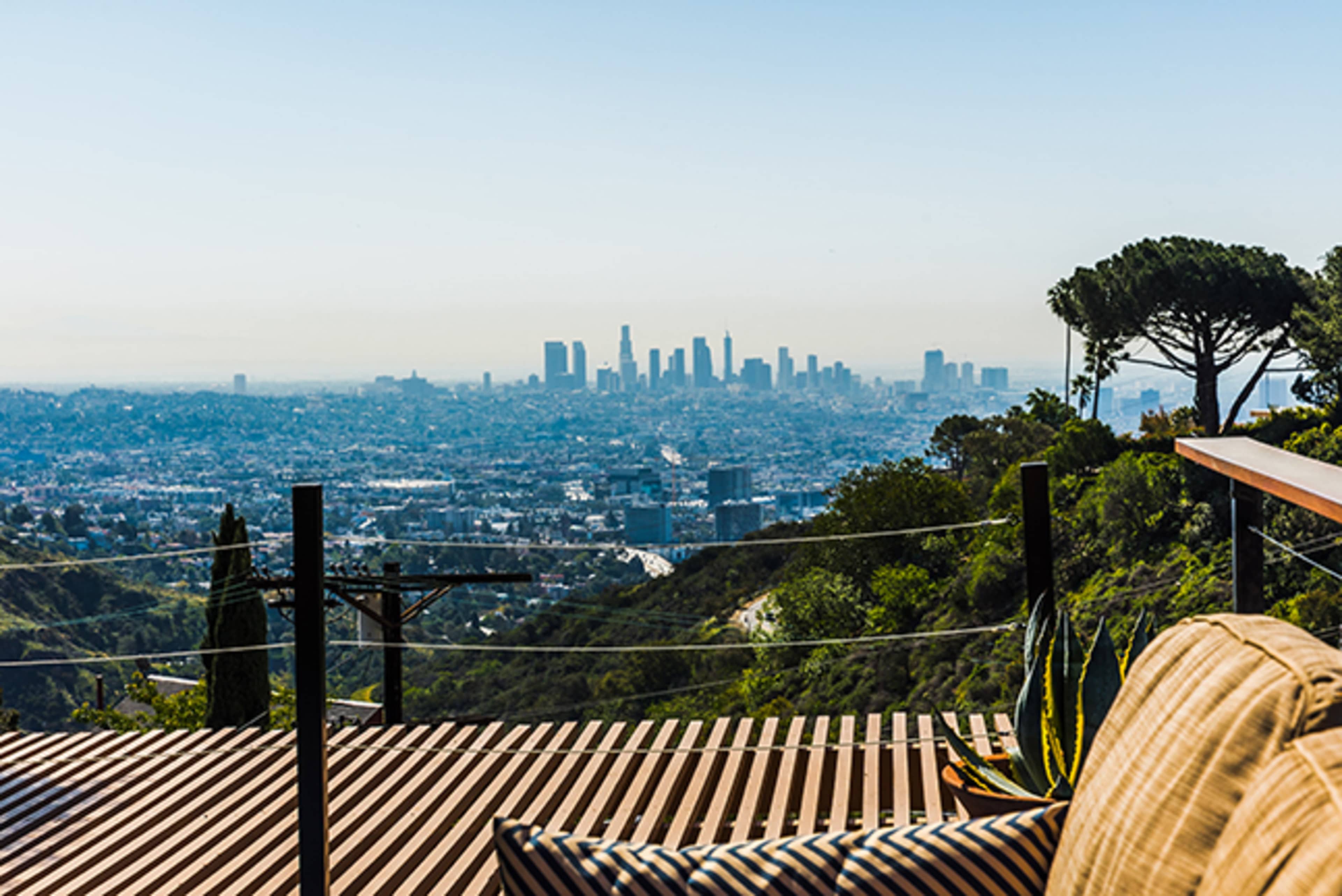 The image shows a view of the Los Angeles skyline set against a clear sky, with a foreground of outdoor seating and greenery.