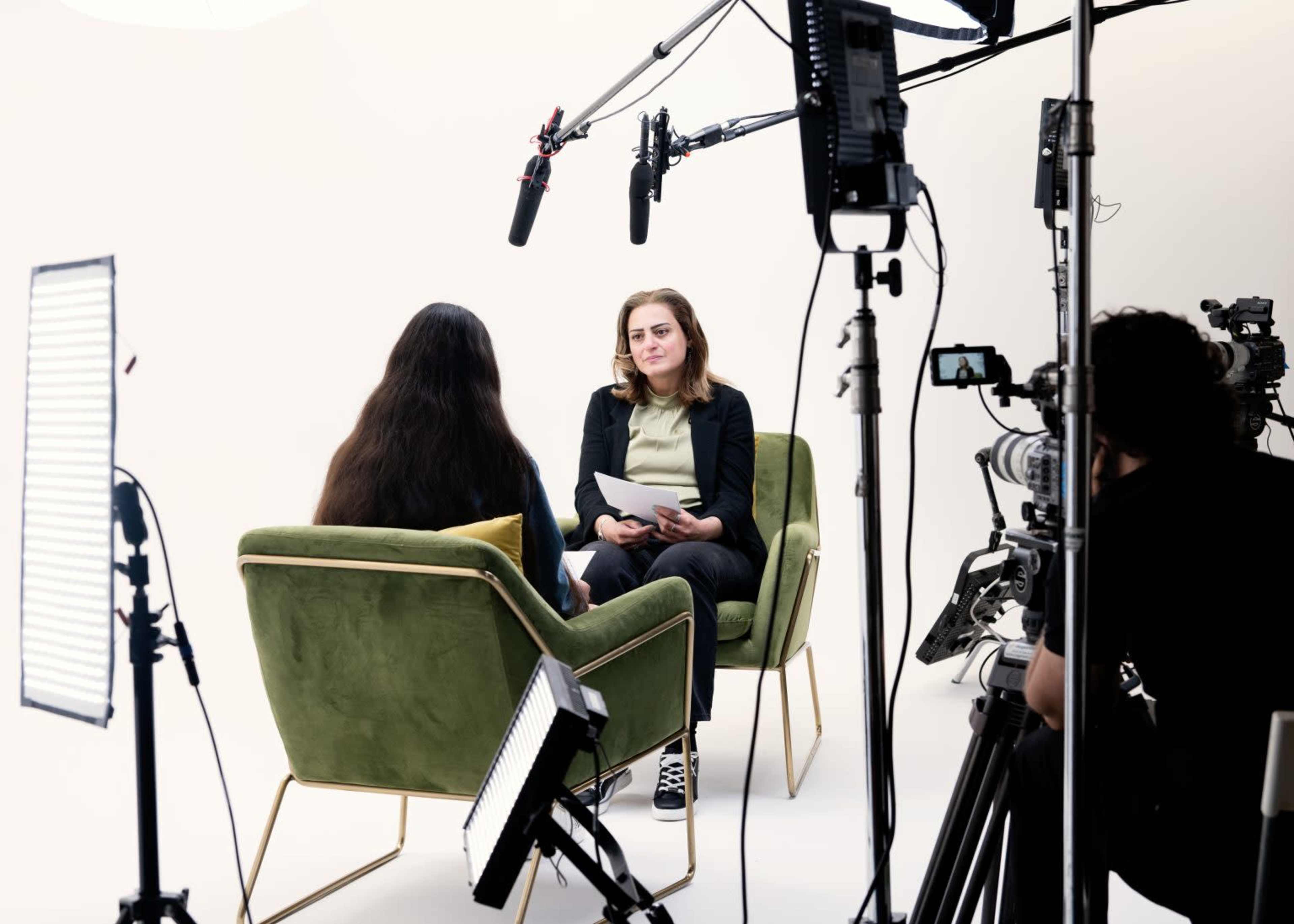 Two individuals are seated in green chairs facing each other in a studio setting, surrounded by film equipment and lighting.