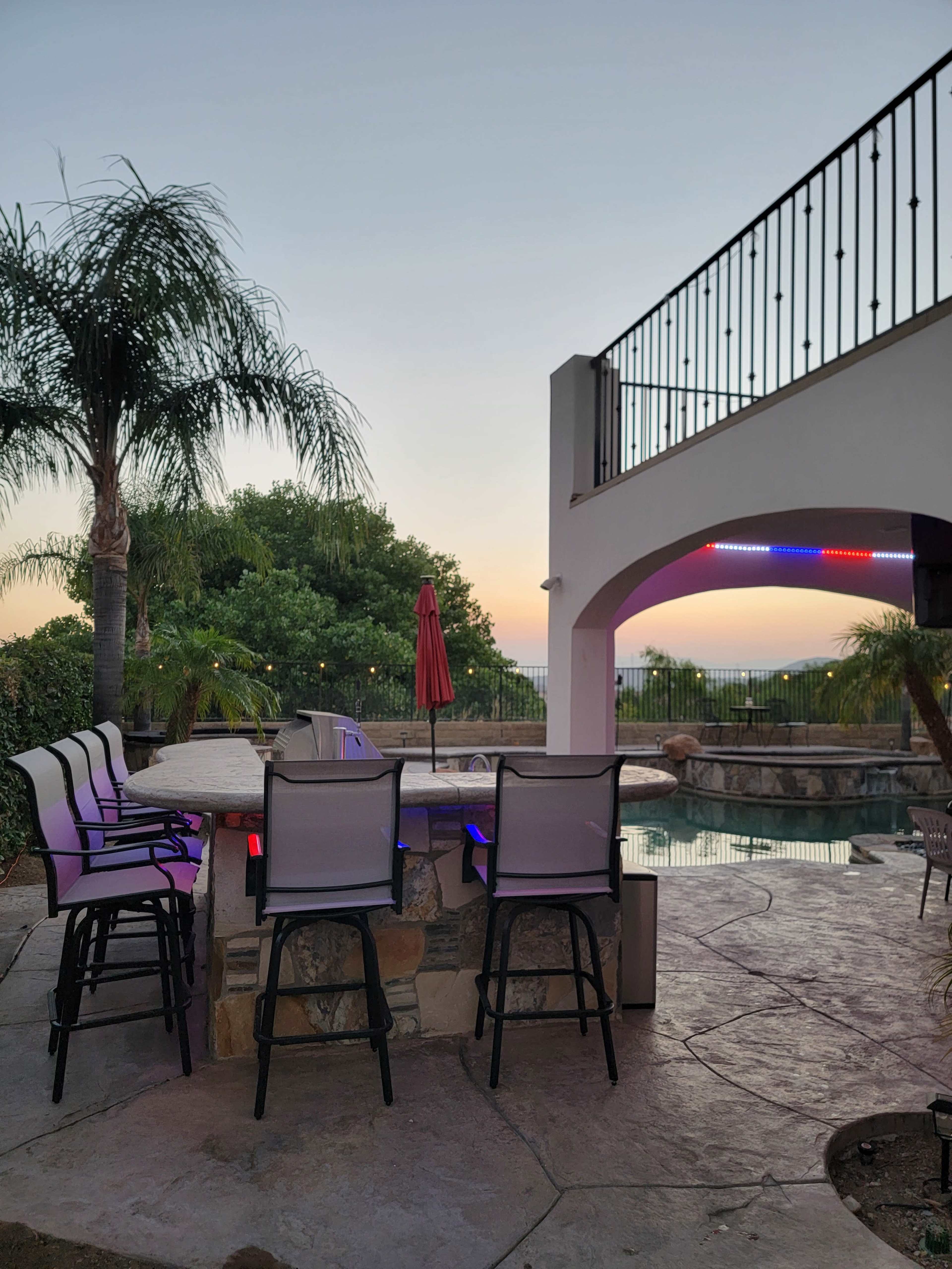 A backyard with bar stools surrounding a stone bar near a swimming pool, illuminated by lights, with palm trees and a sunset in the background.