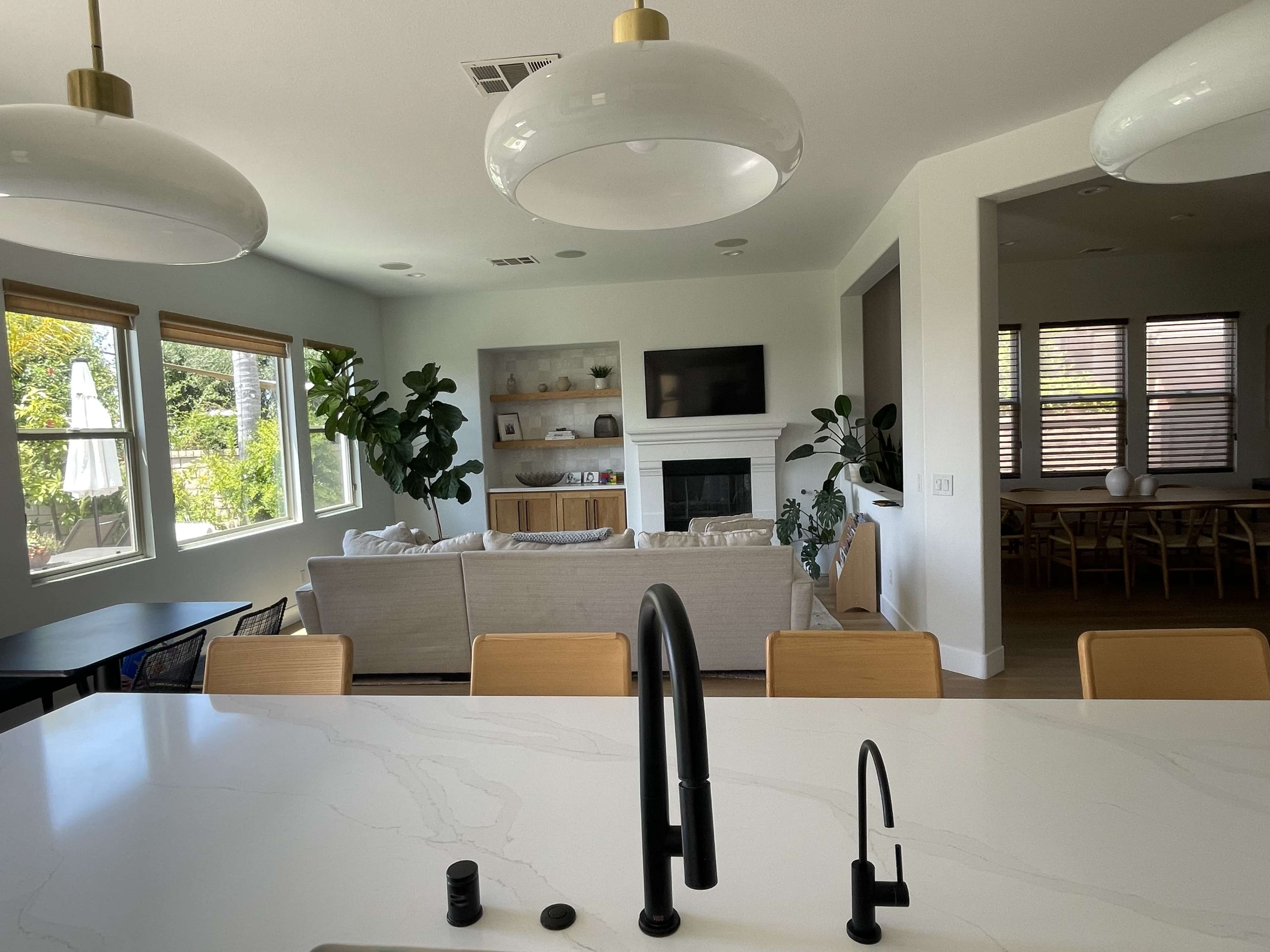 A modern living room features a sectional sofa, a fireplace, and large windows allowing natural light, visible from a kitchen island with black fixtures.