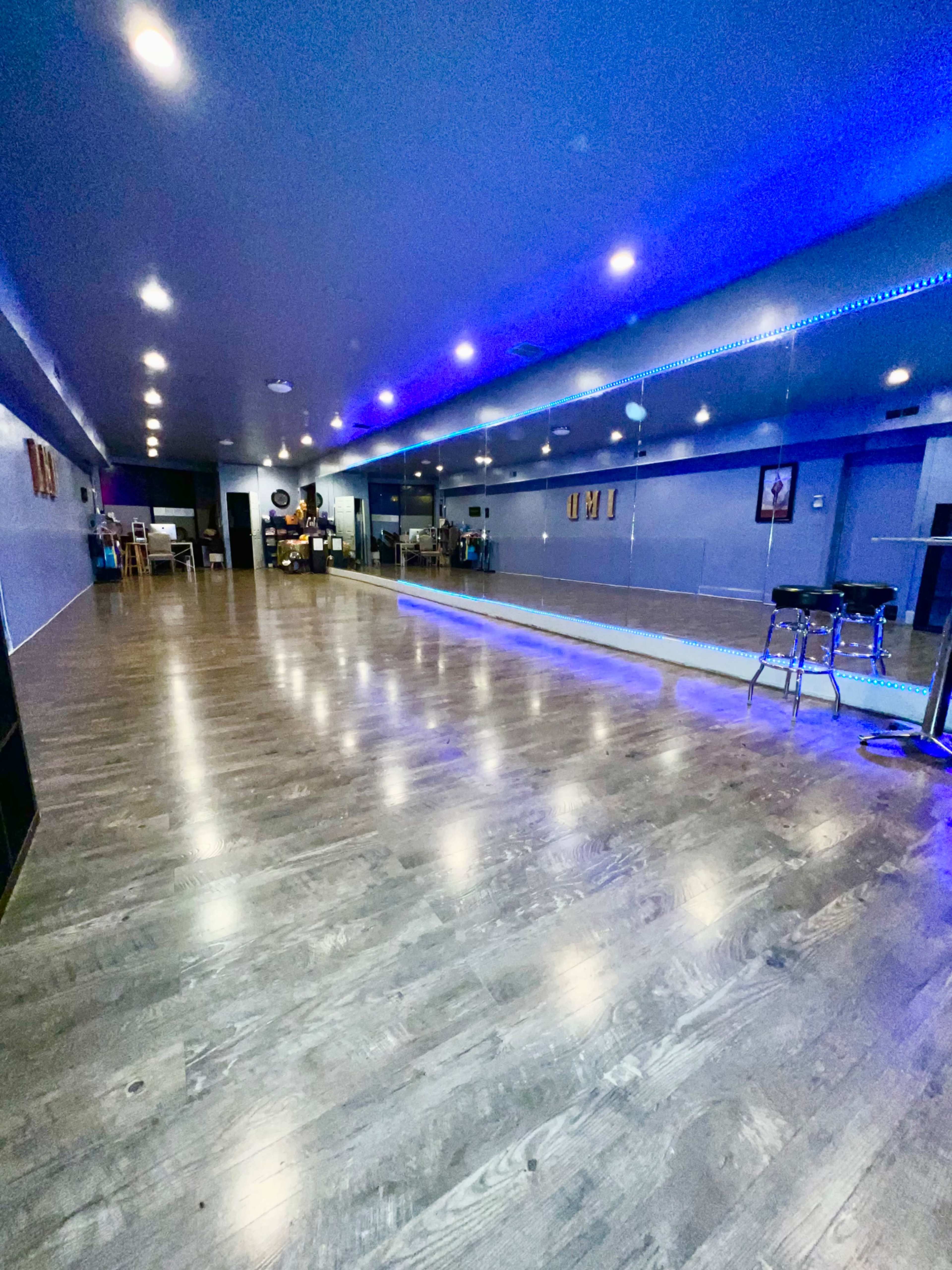 A spacious dance studio with wooden flooring, blue-lit walls, and mirrors reflecting the room's interior.