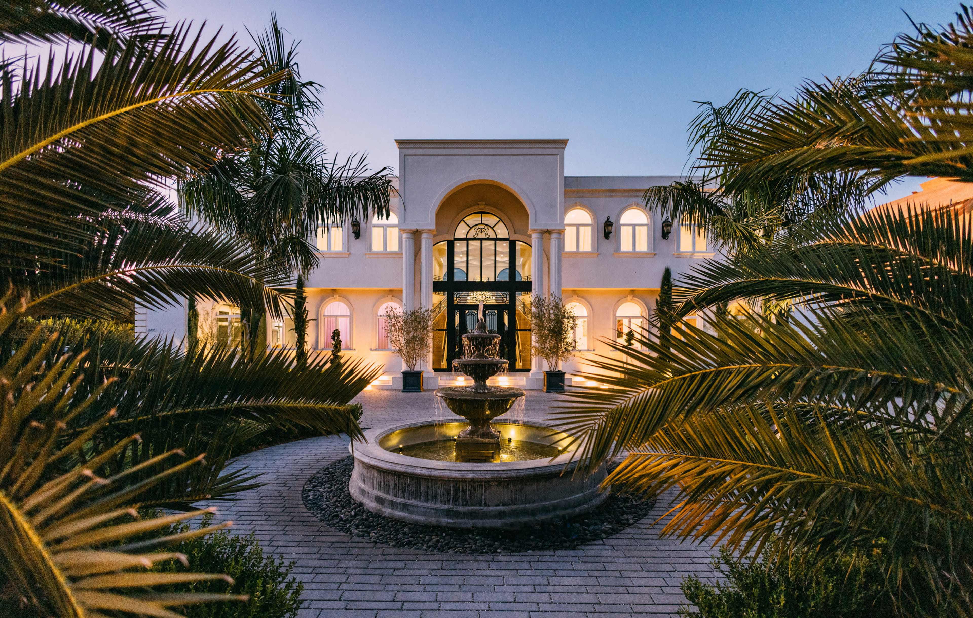 A grand building with an entrance framed by palm trees and a circular fountain in the foreground, illuminated at twilight.