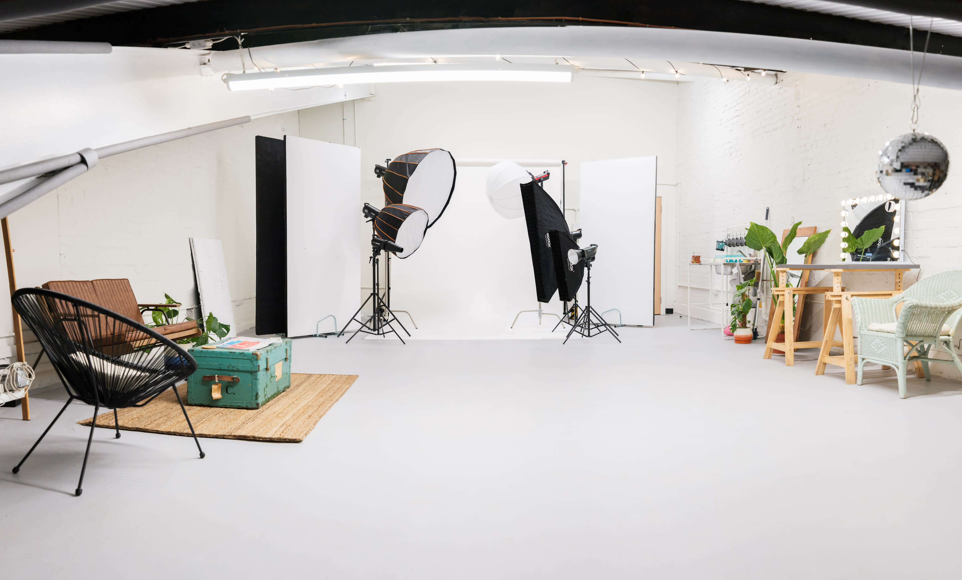 A bright, spacious photography studio features white backdrops, lighting equipment, and various furniture pieces, including a green crate and potted plants.