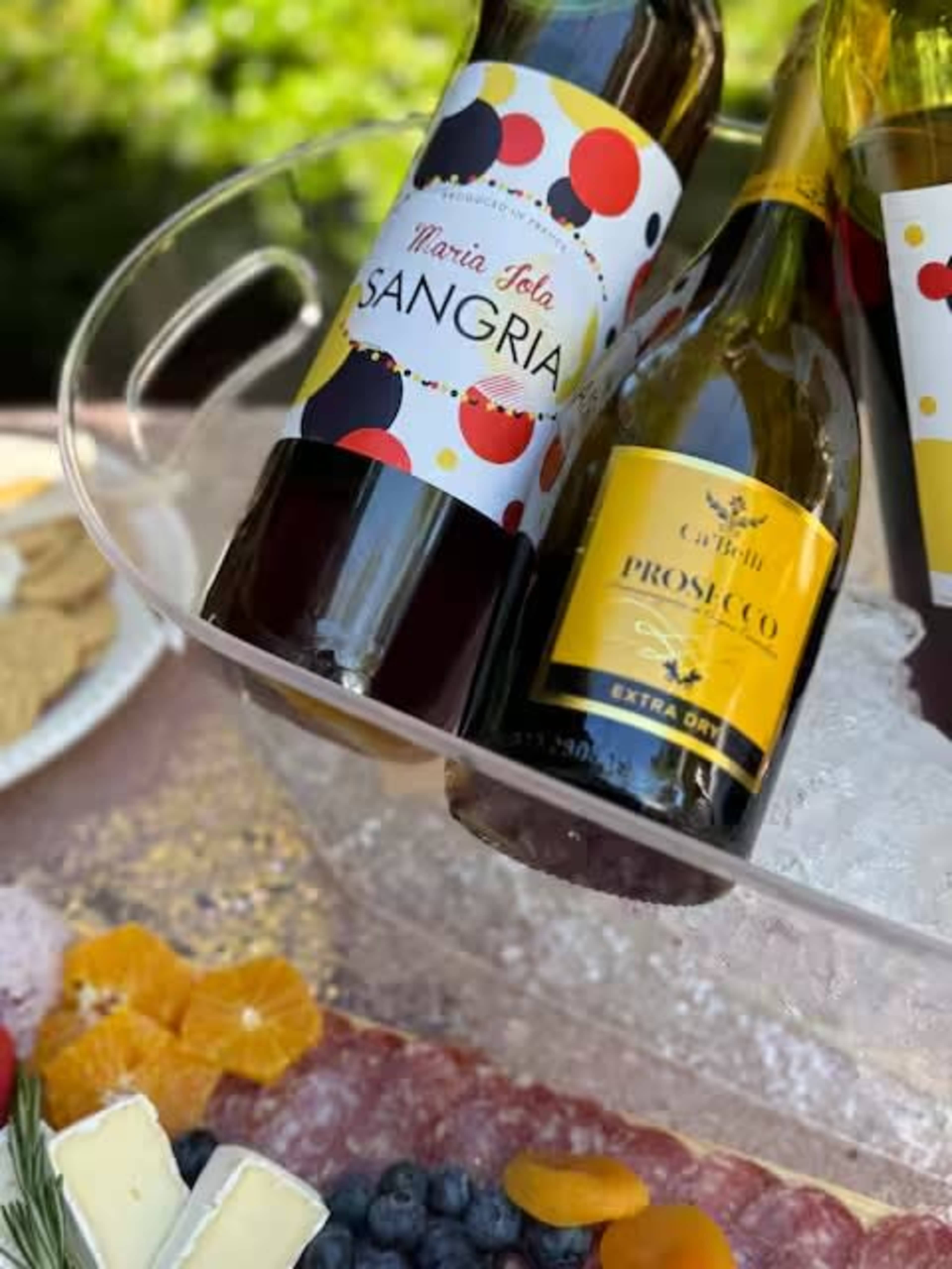 The image shows two bottles of sangria and prosecco chilling in a glass bowl filled with ice, surrounded by an array of snacks, including cheese, fruits, and dried fruits.