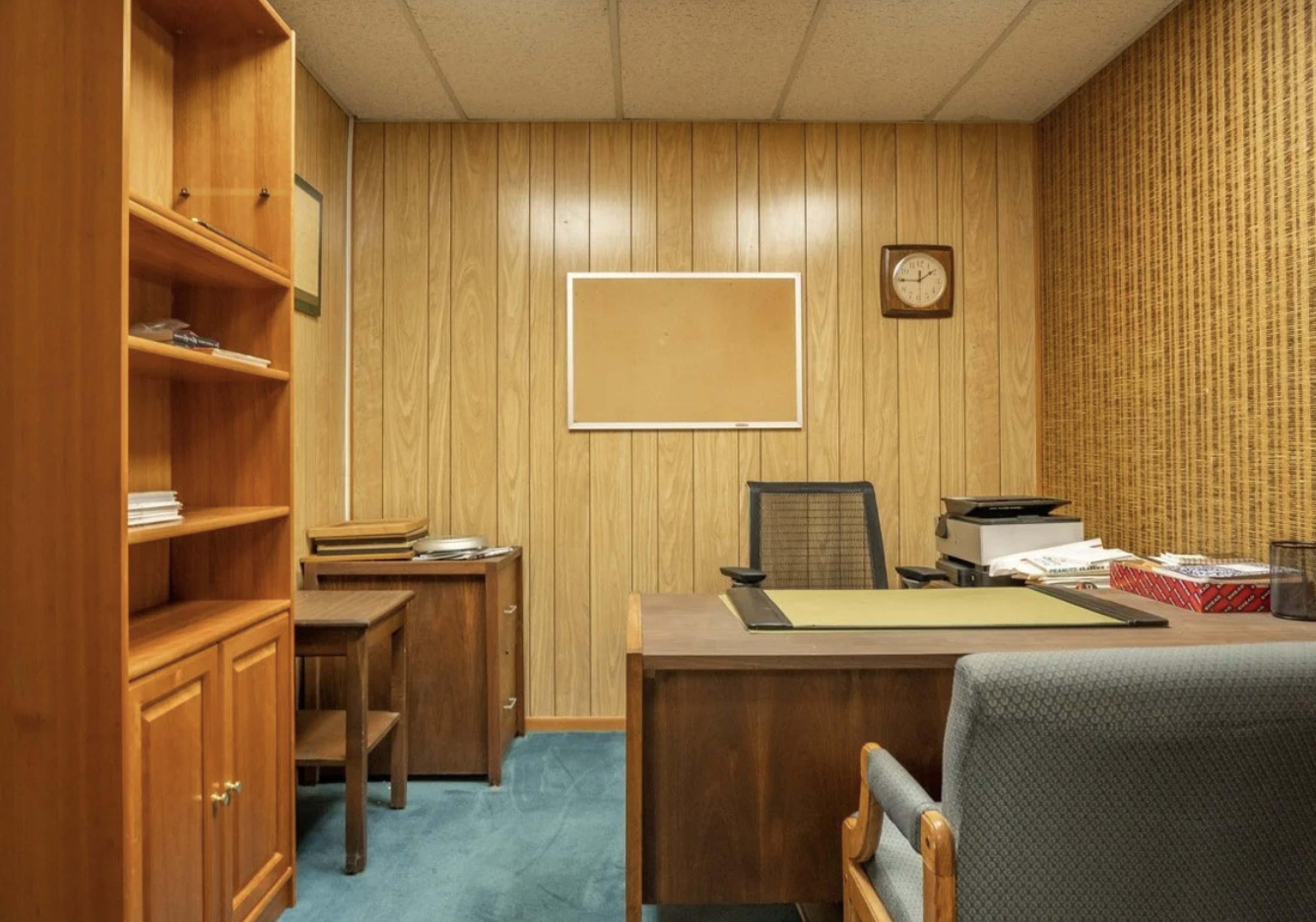The image shows a small office with wooden paneling, a desk with a green mat, a chair, a bookshelf, and a clock on the wall.