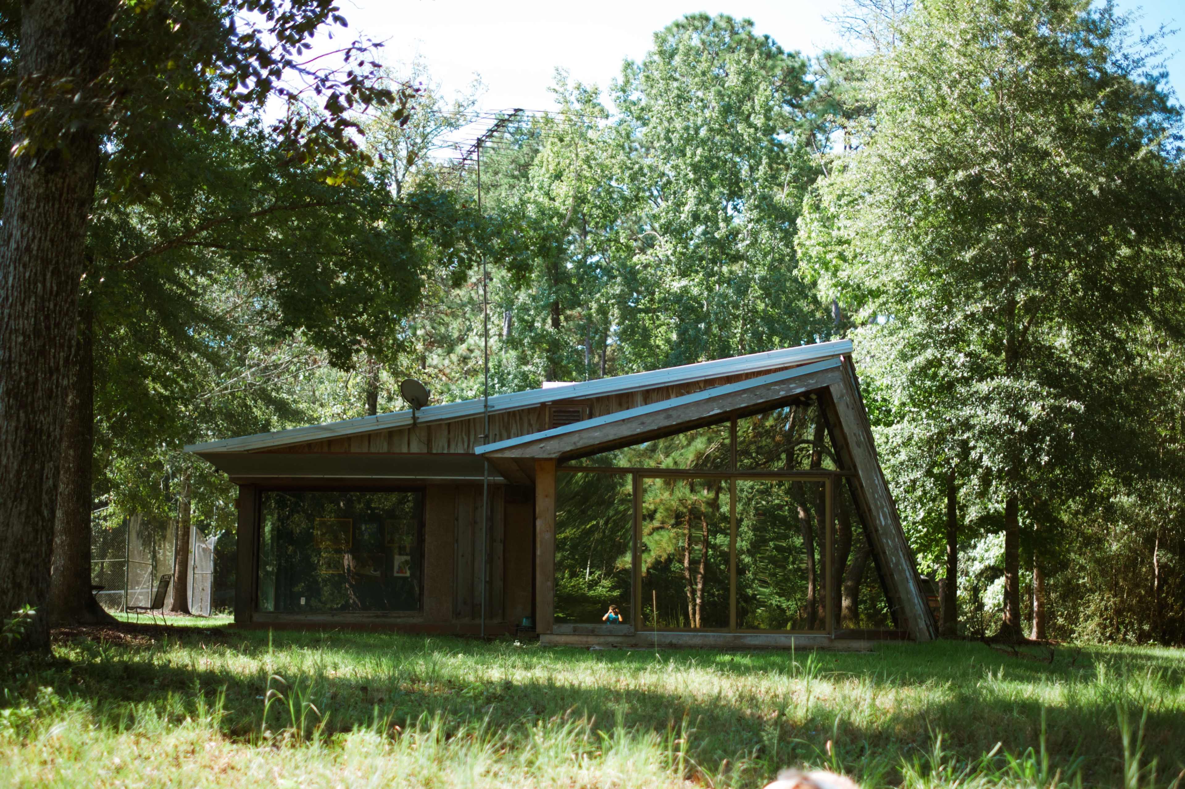 A modern house with large glass windows and a slanted roof is situated among trees in a grassy area.