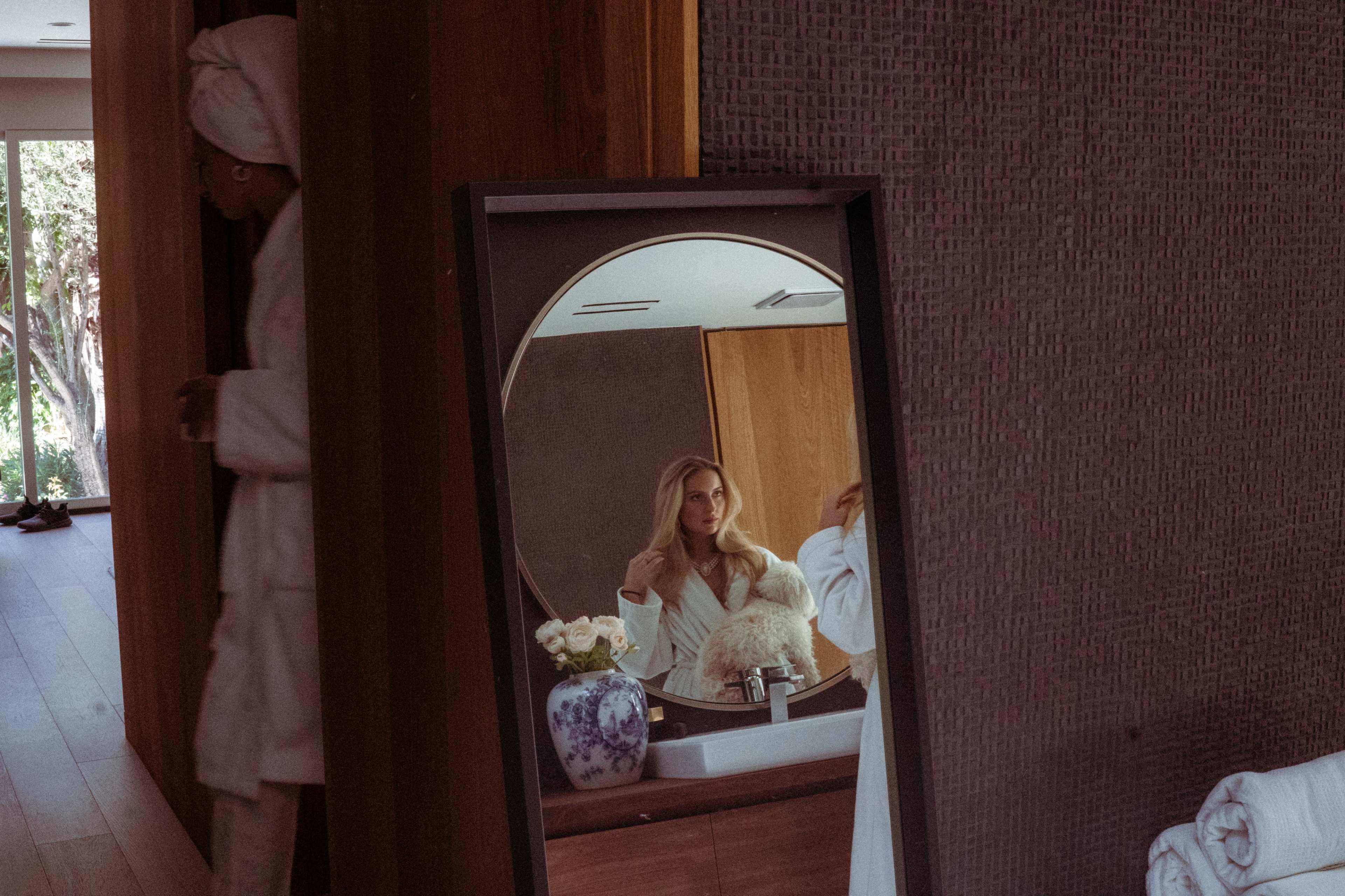 A woman in a bathrobe stands next to a large mirror reflecting her image and a vase with flowers on a bathroom counter.