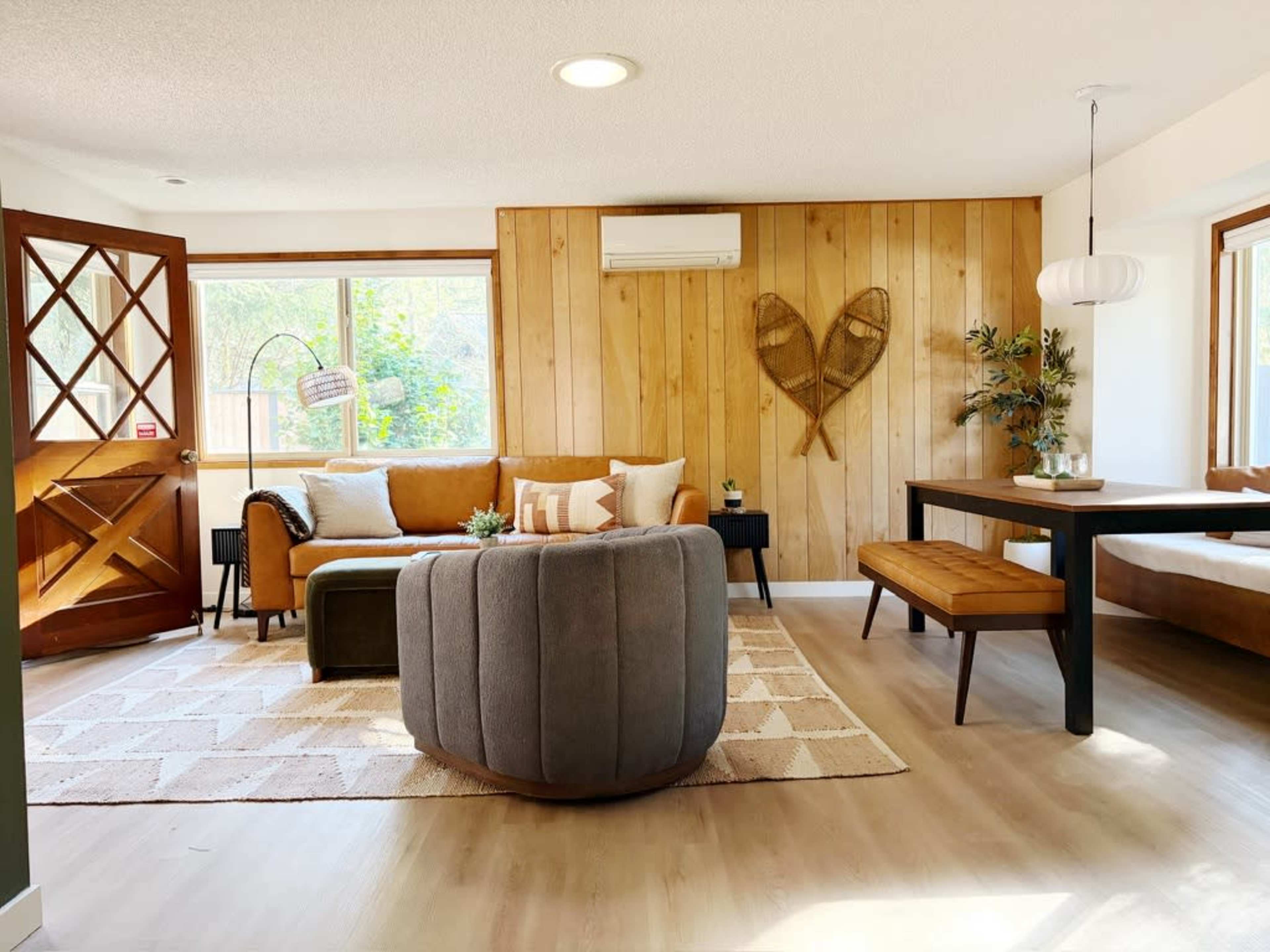 The image shows a cozy living space with a brown sofa, a round gray ottoman, a wooden dining table, and decor elements like plants and wall art.