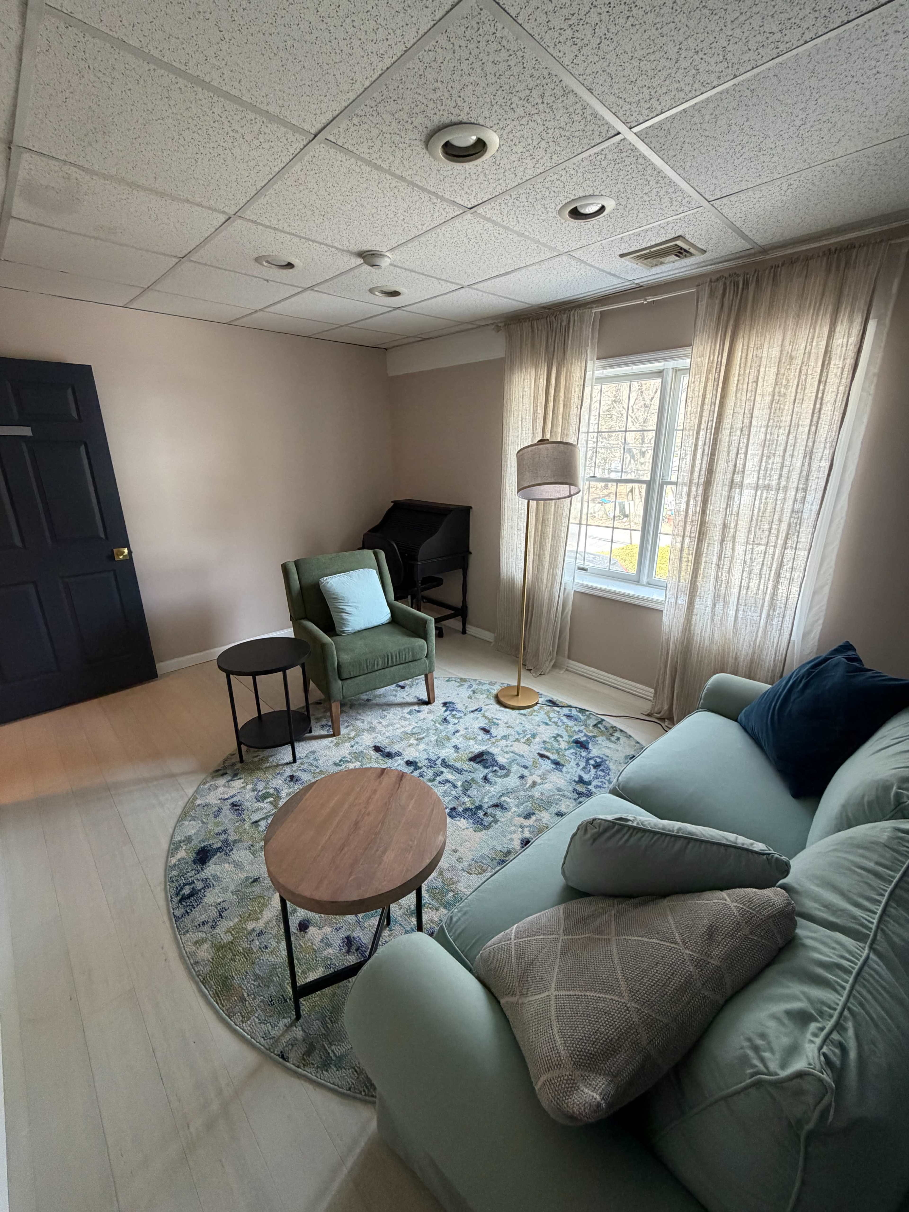 The image depicts a small living room featuring a light green sofa, a green armchair, a round wooden coffee table, a floor lamp, and large windows with sheer curtains.