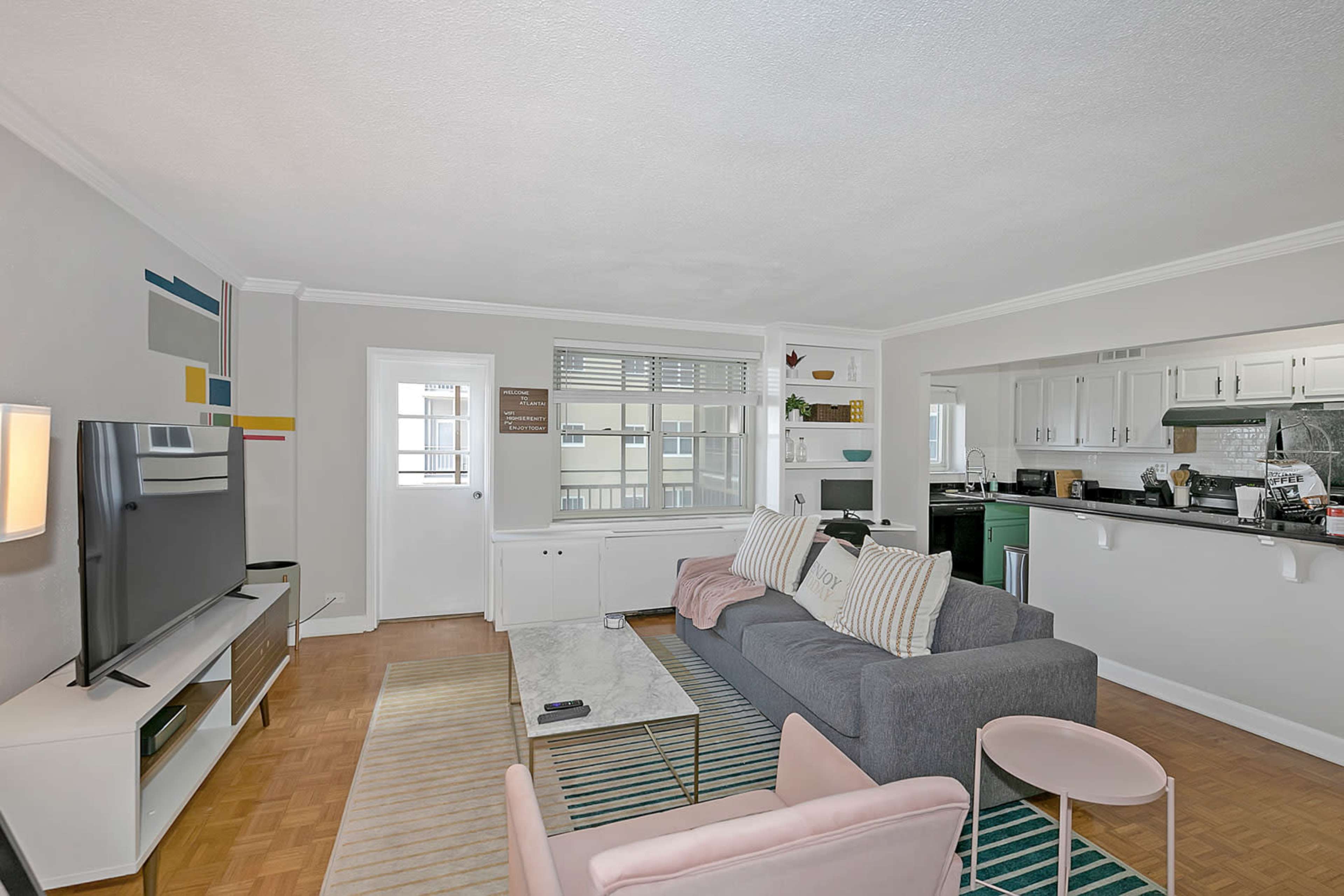 The image shows a modern living room featuring a gray sofa, a light pink armchair, a patterned rug, and a television on a white media console, with a view into a kitchen area.