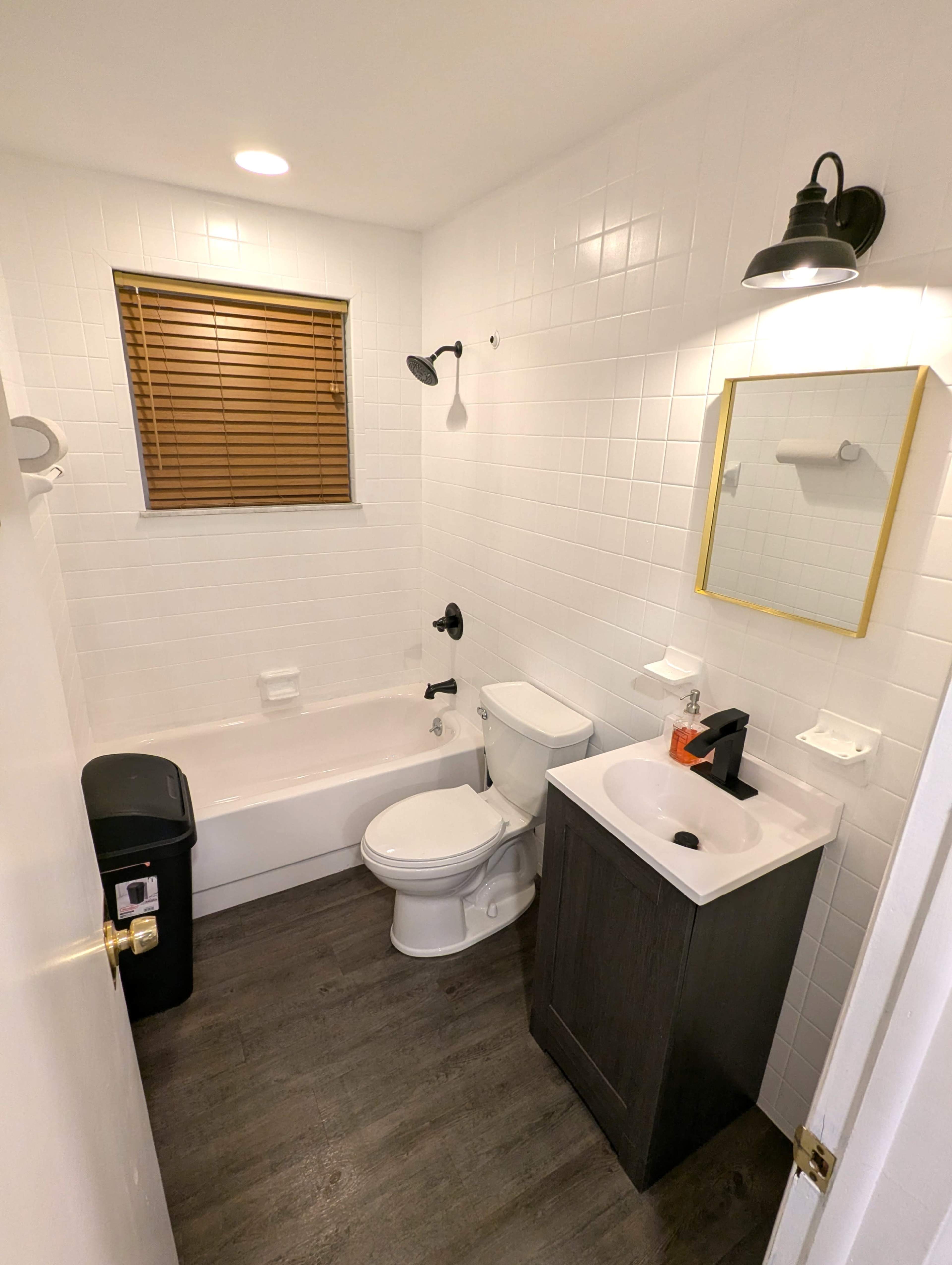 The image shows a compact bathroom featuring a bathtub with a shower, a toilet, a sink with a dark cabinet, and a window with wooden blinds.