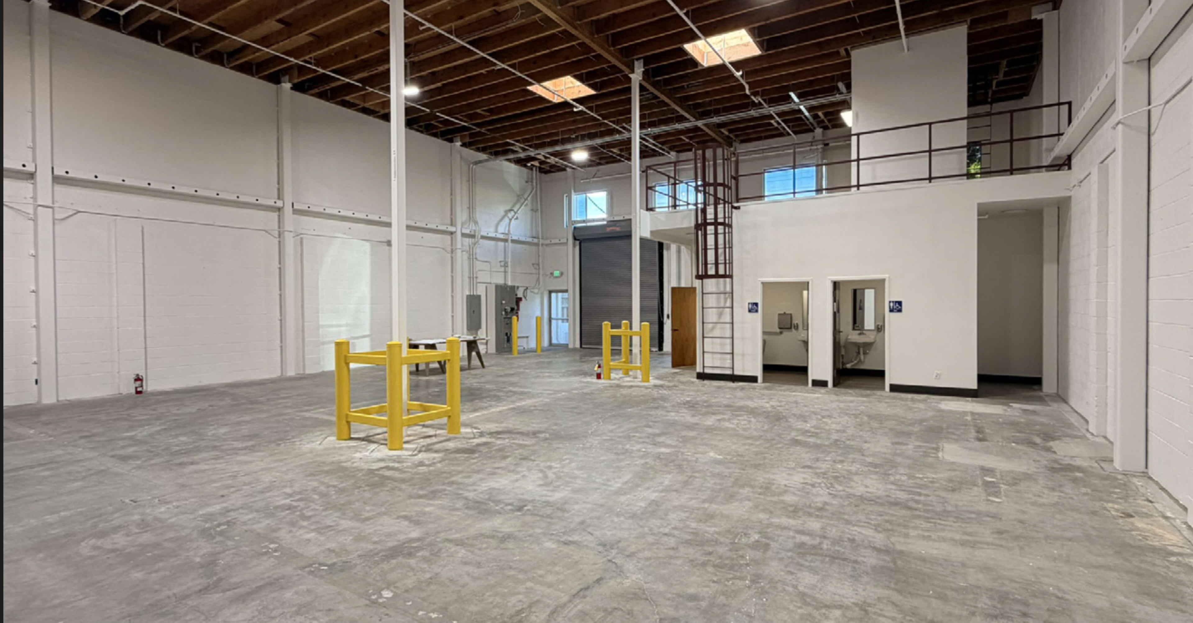The image shows an empty industrial space with concrete flooring, white walls, and yellow safety barriers, featuring a restroom area and a staircase in the background.