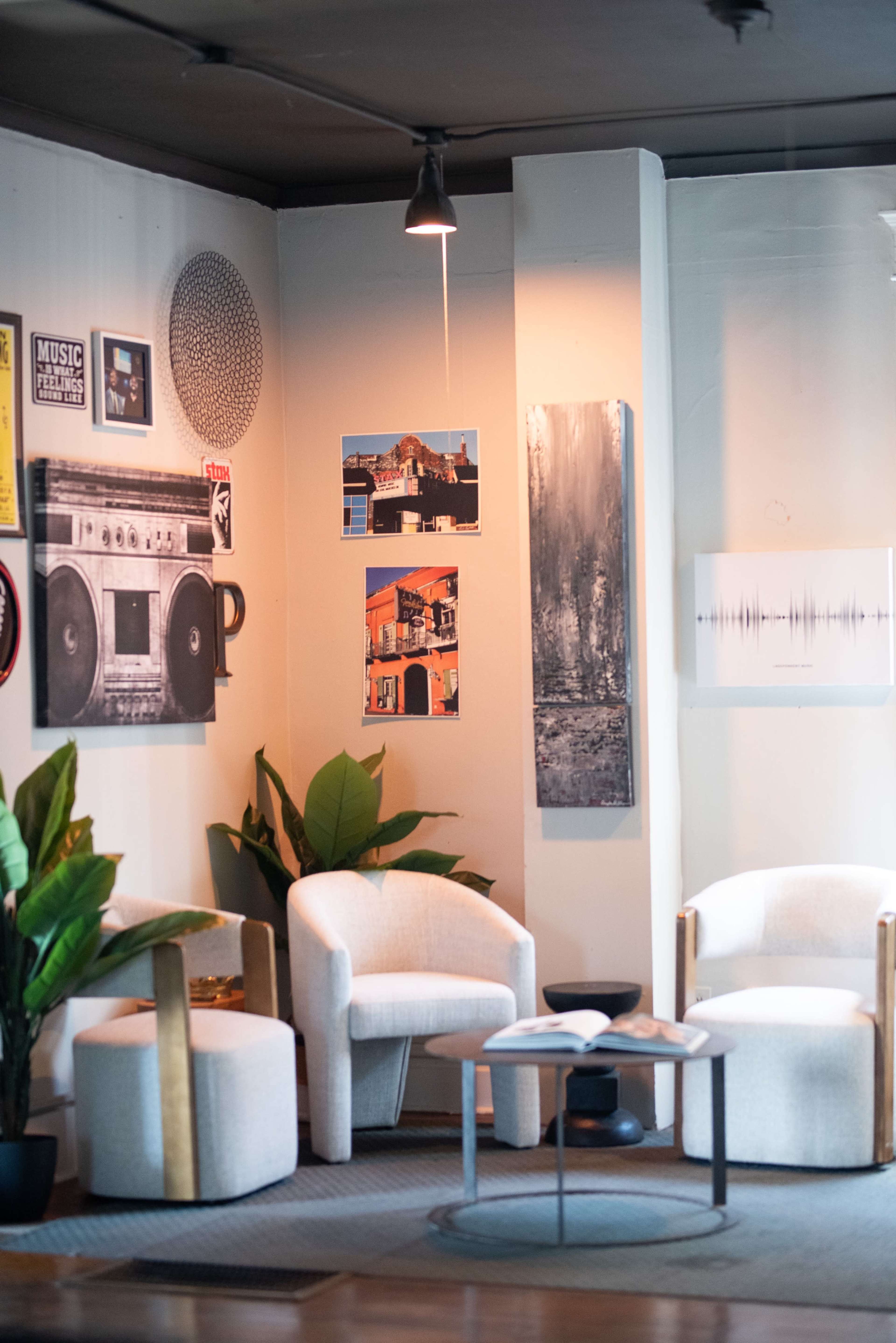 The image shows a modern seating area with two white chairs, a small table, and a potted plant, adorned with various framed artworks on the walls.