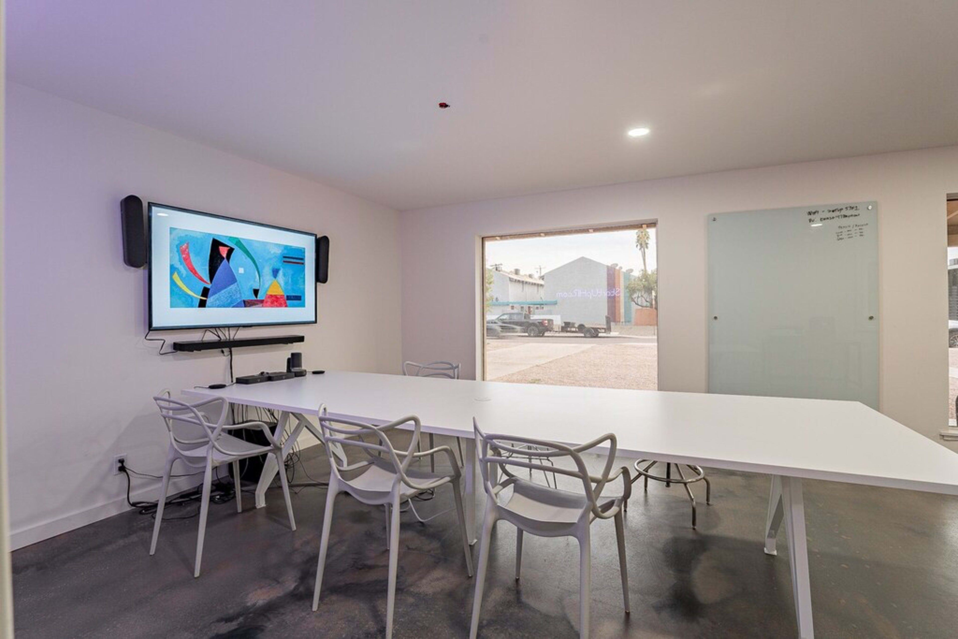 A modern conference room features a white table with six transparent chairs, a wall-mounted screen displaying colorful artwork, and a large window overlooking an outdoor area.