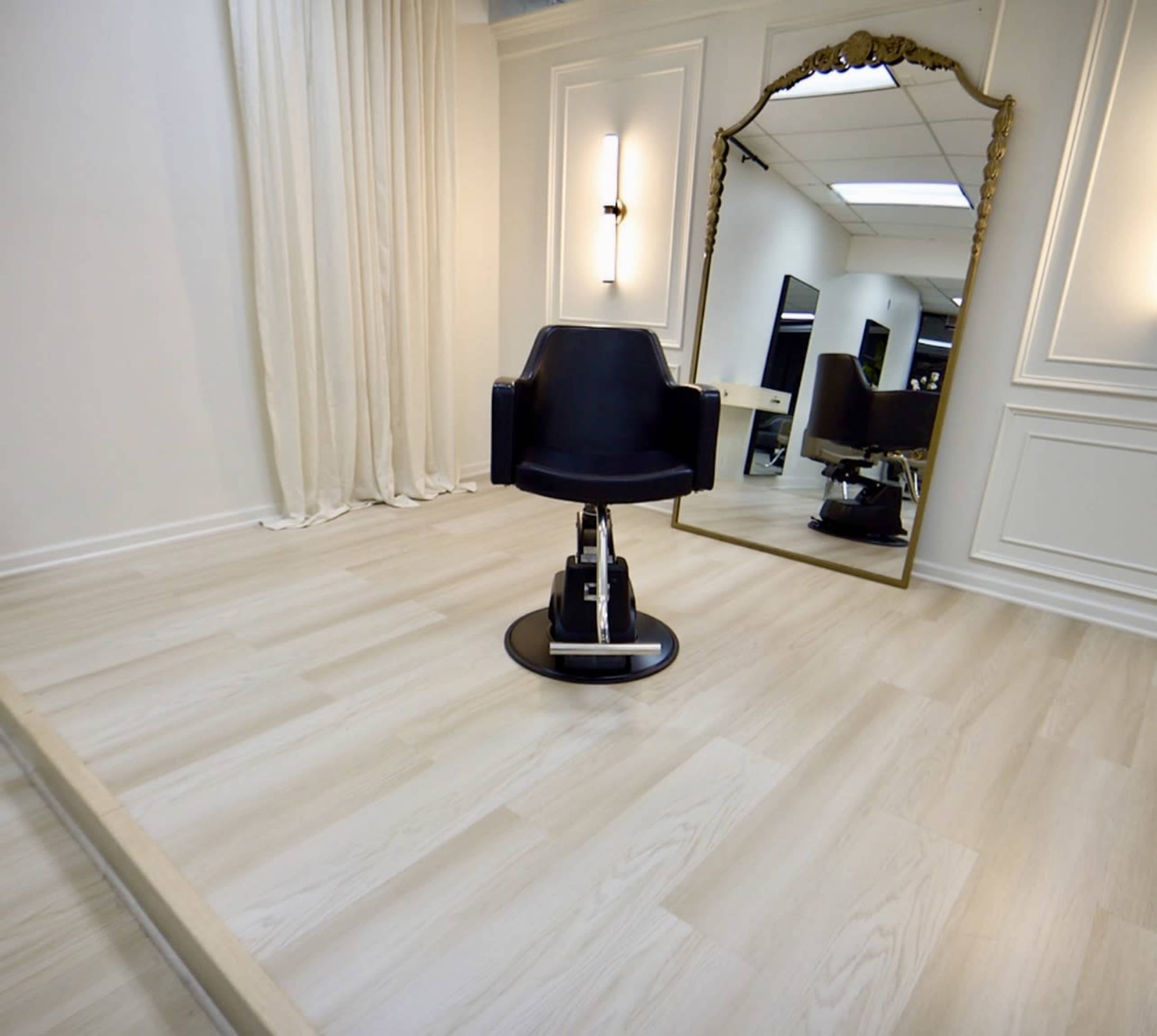 A black styling chair is positioned on a light-colored wooden floor, facing a large mirror in a softly lit, minimalistic salon space.