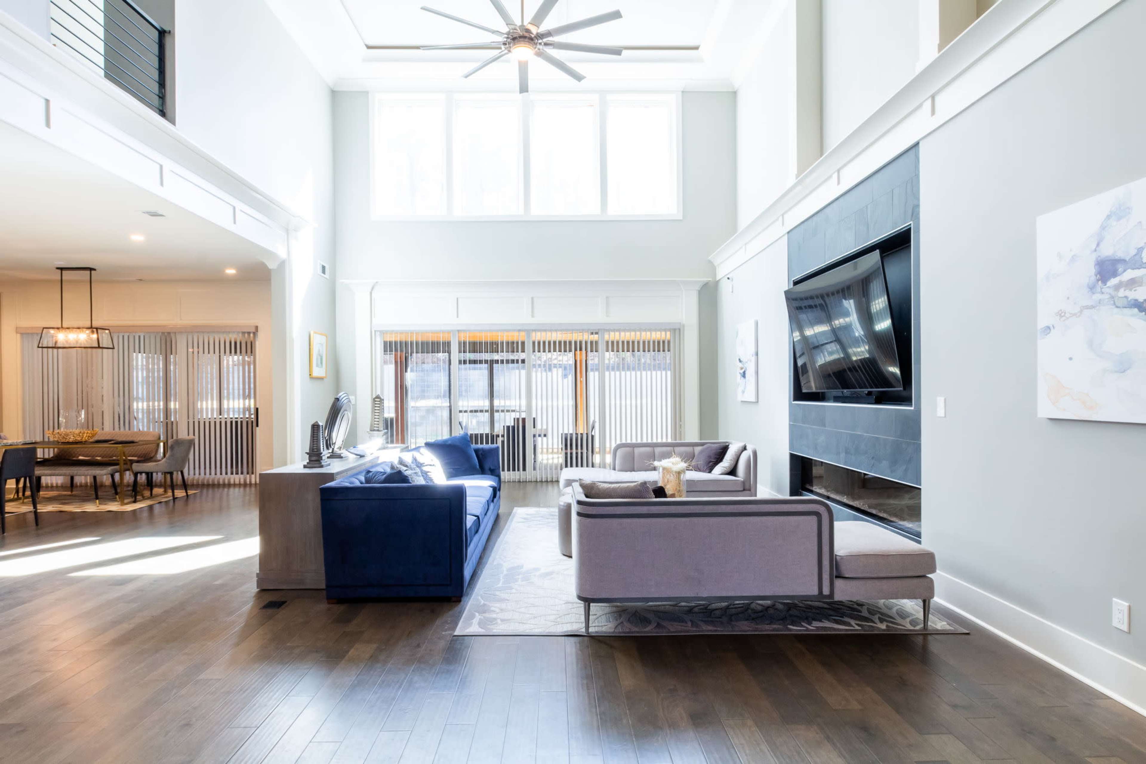 The spacious living area features a blue sofa and a gray sectional, with large windows allowing natural light to fill the room.