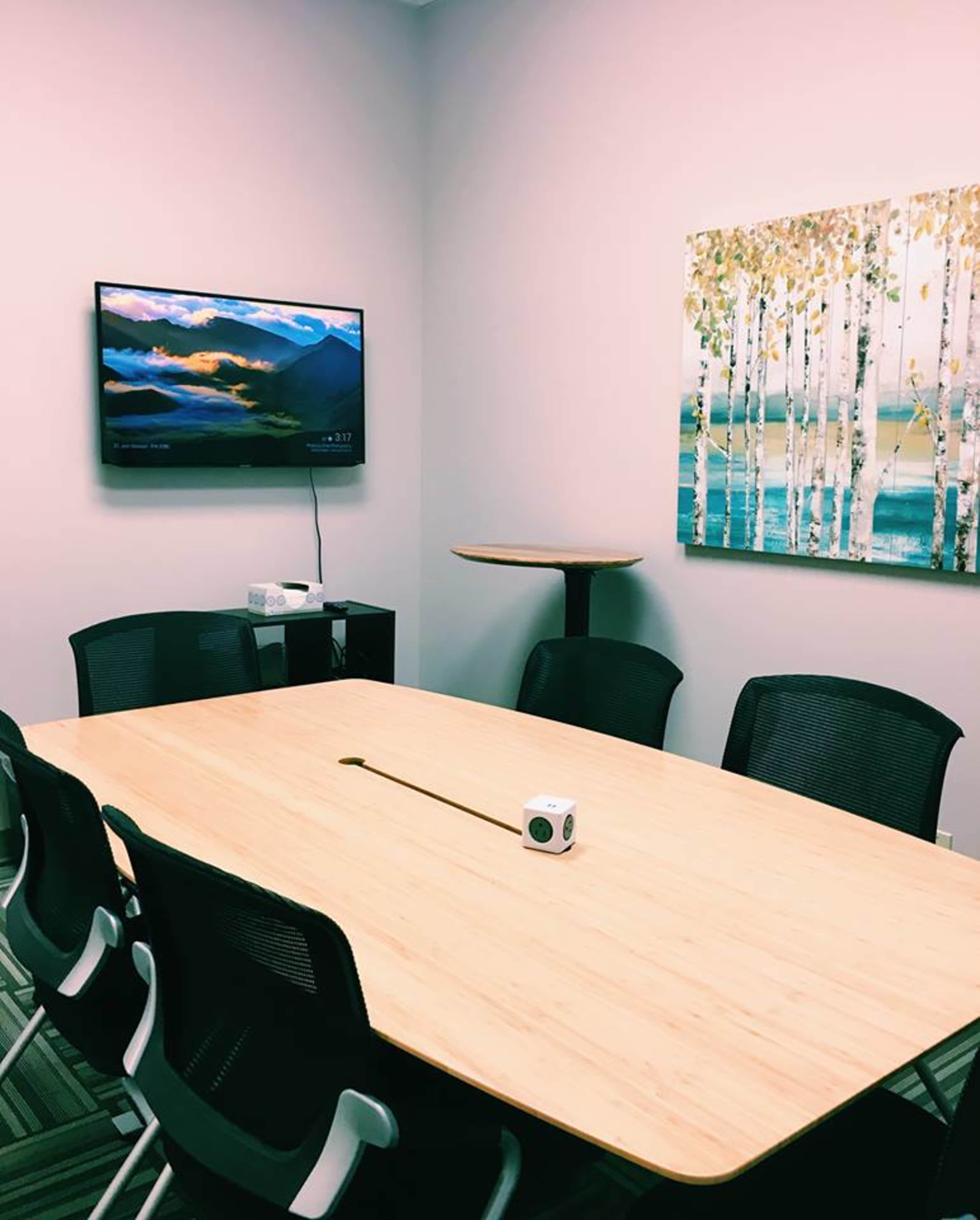A conference room features a large wooden table surrounded by chairs, a wall-mounted television displaying a landscape, and a piece of artwork depicting a forest scene.