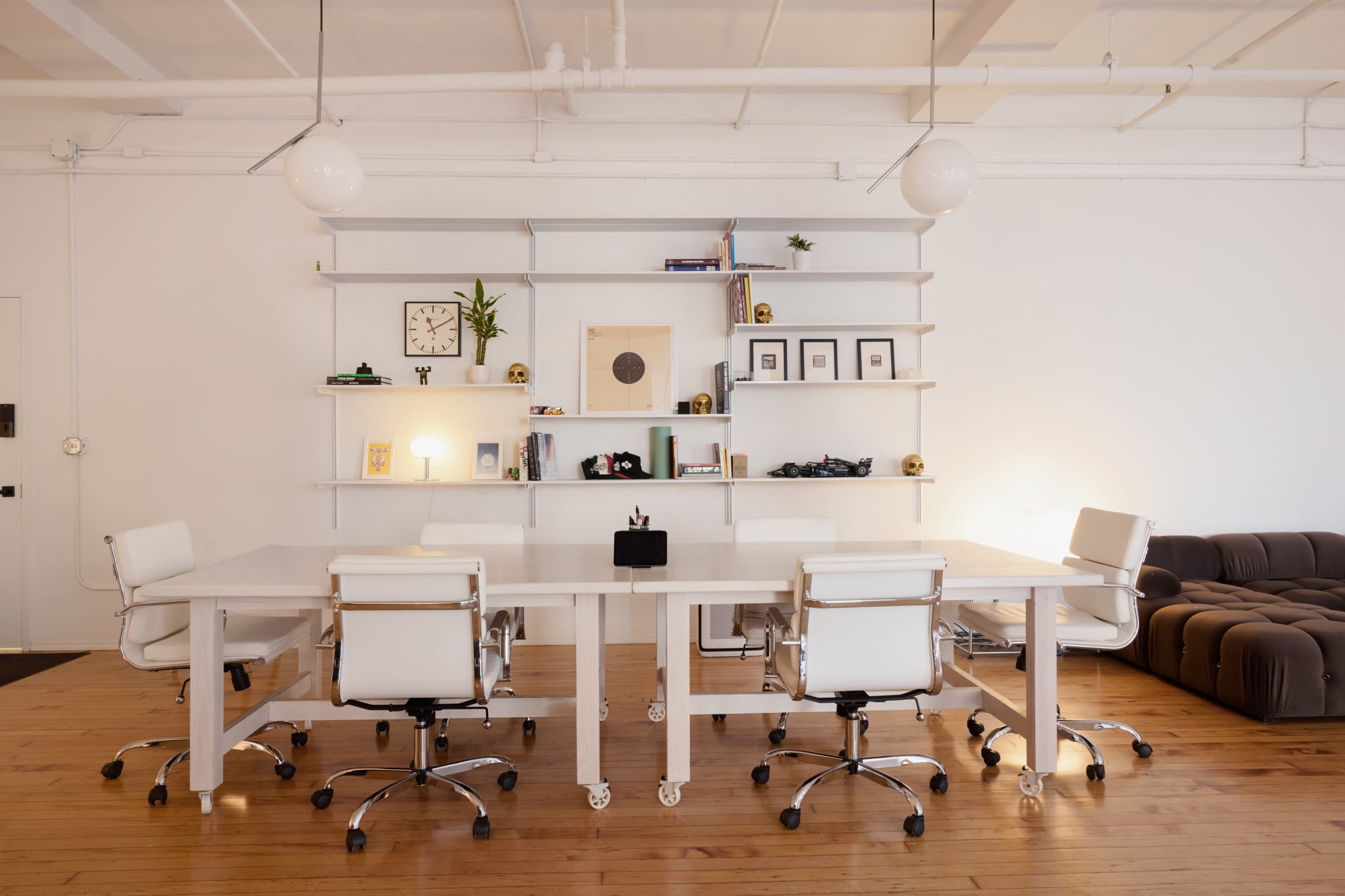 A modern conference room features a large white table surrounded by four white chairs, with shelves displaying various items and framed pictures on the wall.