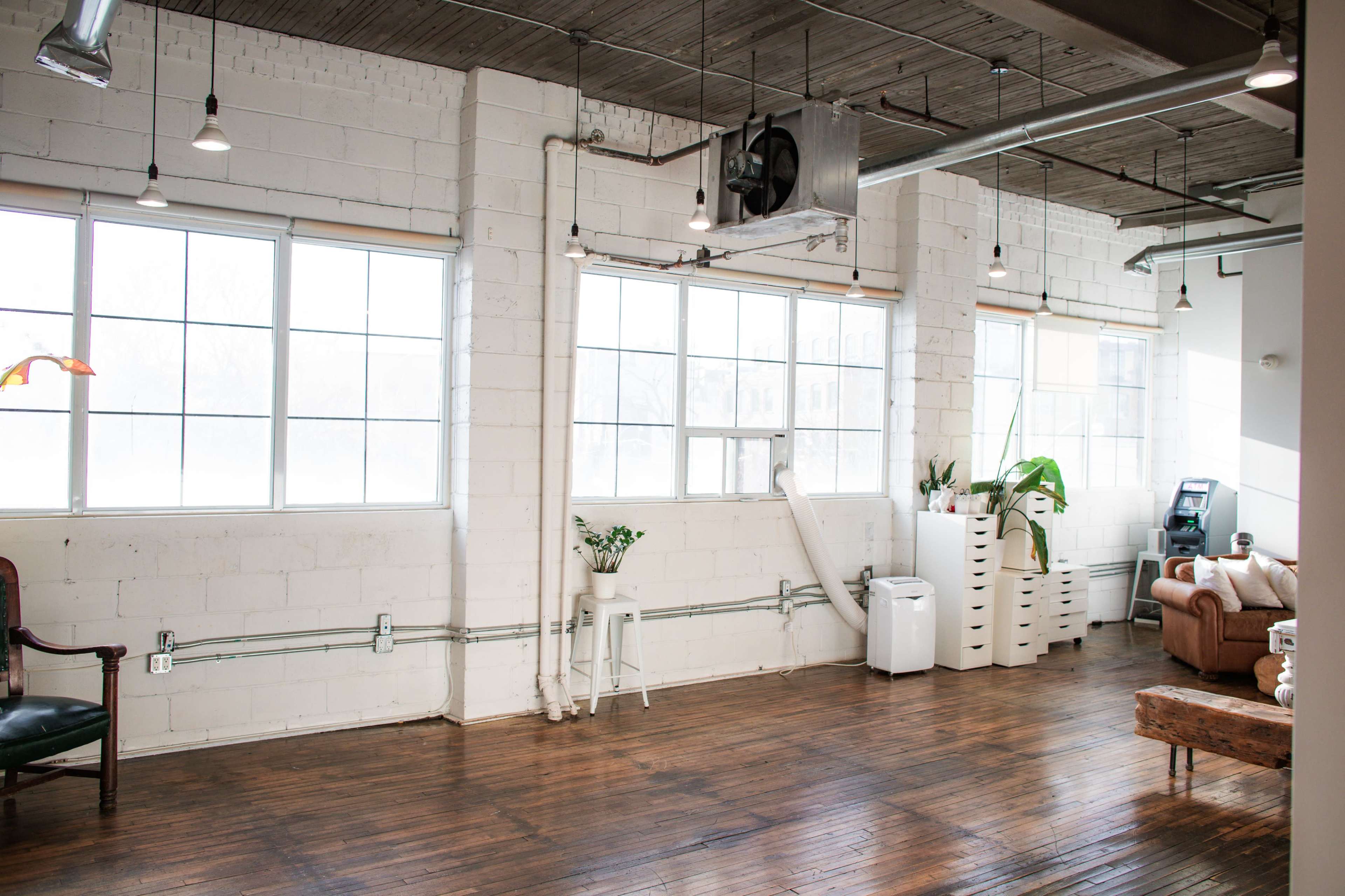 An industrial-style room with large windows, wooden flooring, and minimal furnishings, including a chair, a potted plant, and a couch.