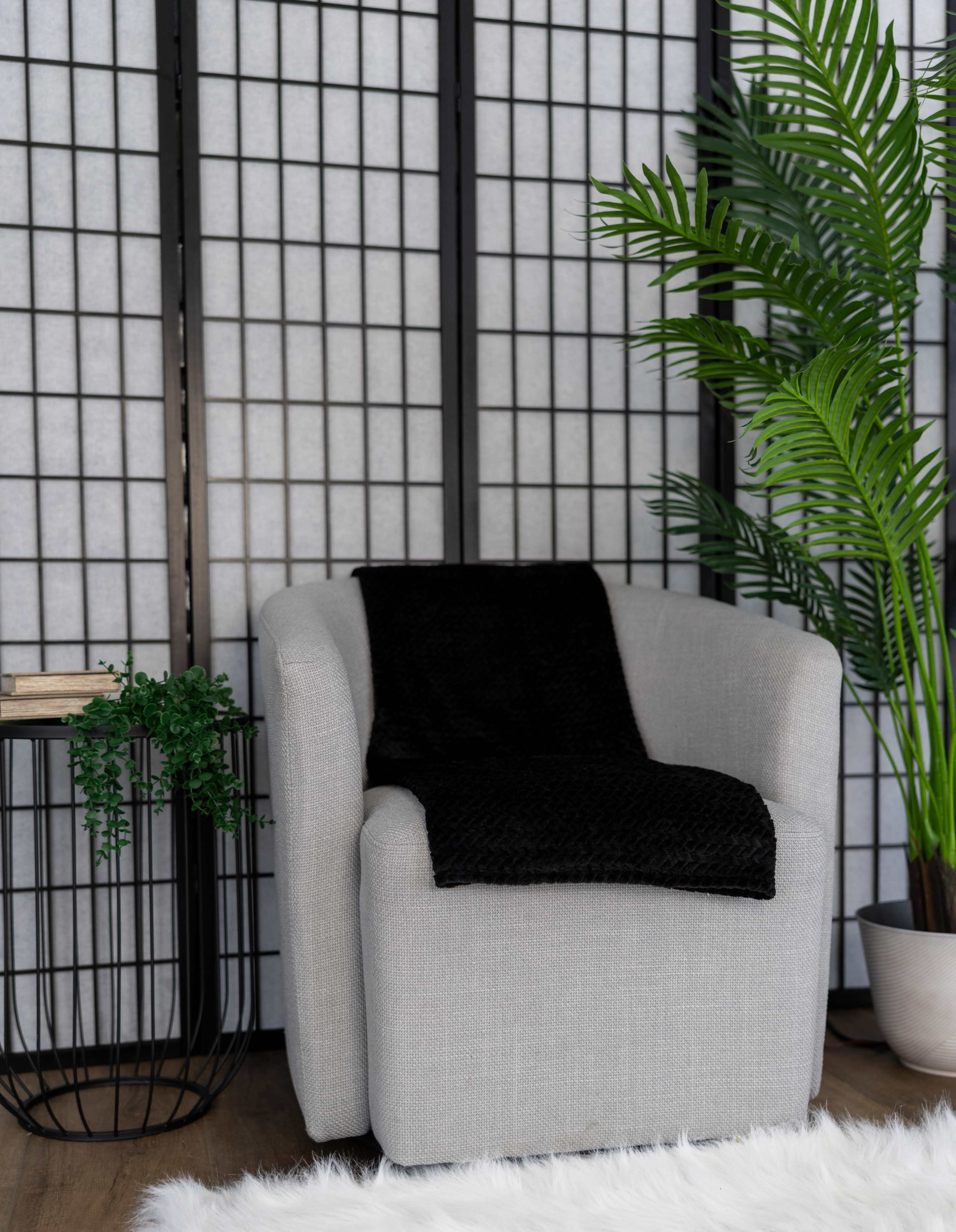 A gray armchair with a black throw blanket is positioned next to a black wire side table with a small plant, set against a backdrop of a wooden floor and a screen of thin vertical slats.