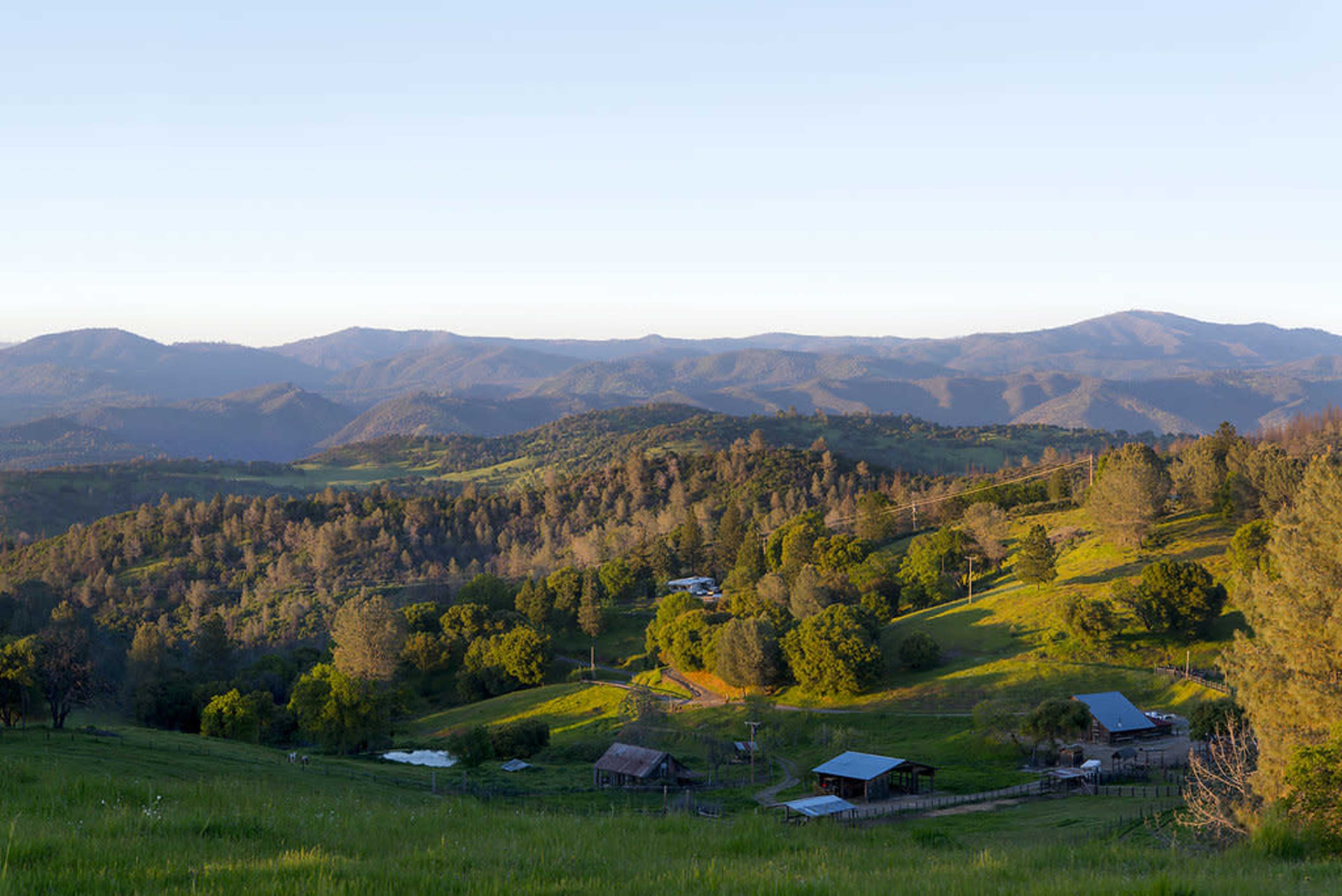 The image shows a sprawling green landscape with gently rolling hills, farm buildings, and a backdrop of distant mountains under a clear sky.