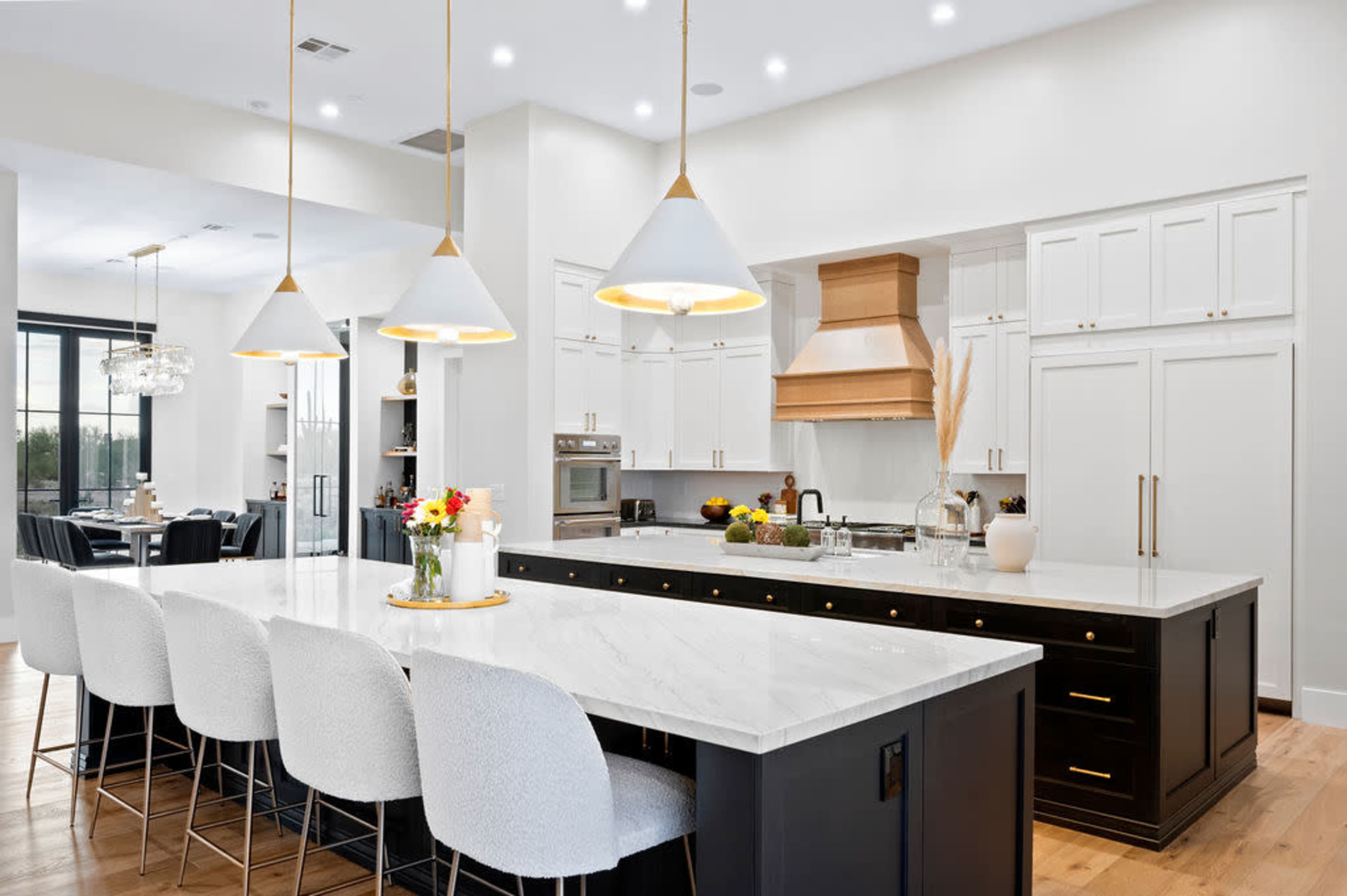 A modern kitchen features a large island with a marble countertop, white bar stools, and a wood-paneled range hood above the stove.